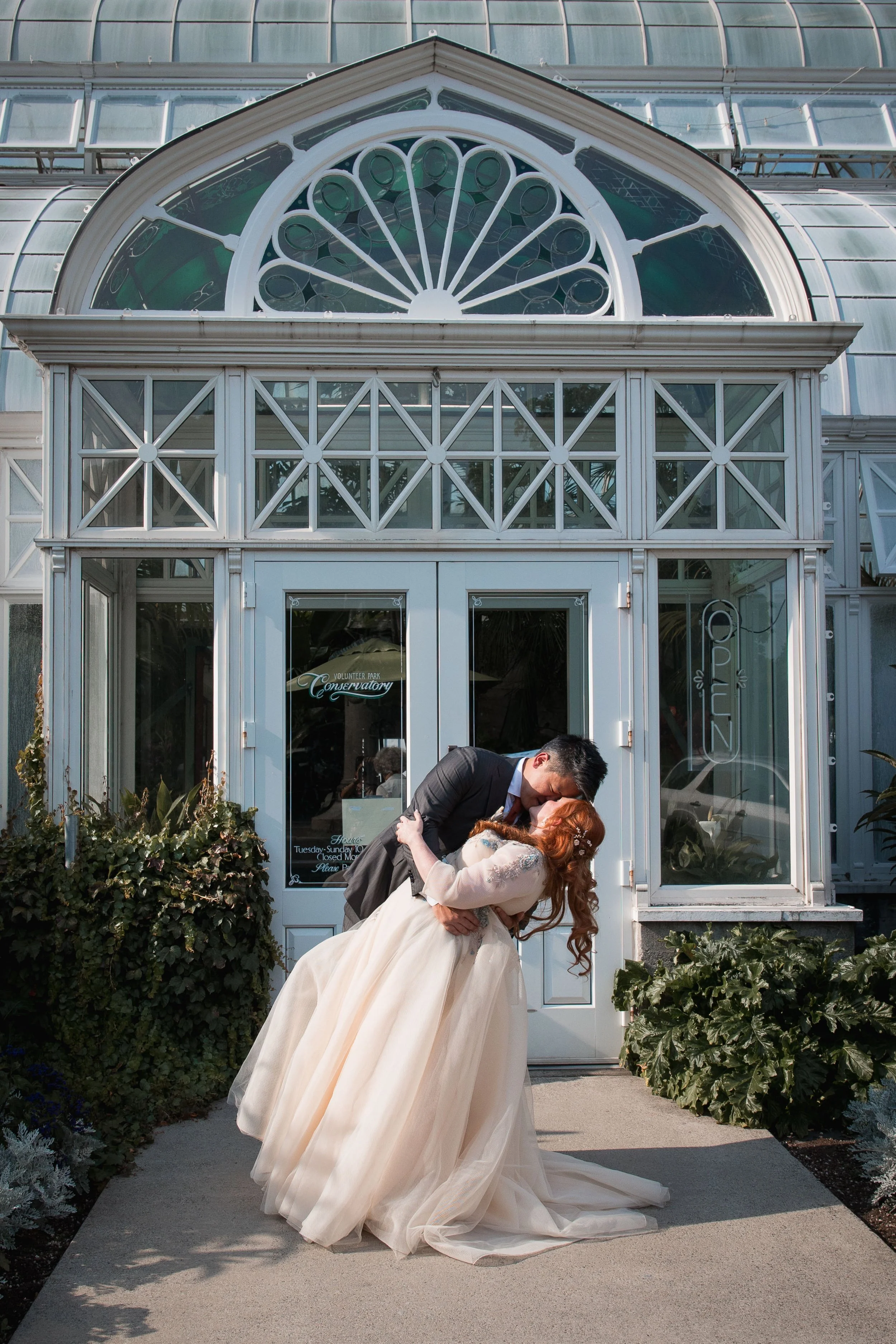 A groom and bride sharing a kiss outside a conservatory during a wedding, with the groom wearing a dark suit and the bride wearing a cream wedding gown.