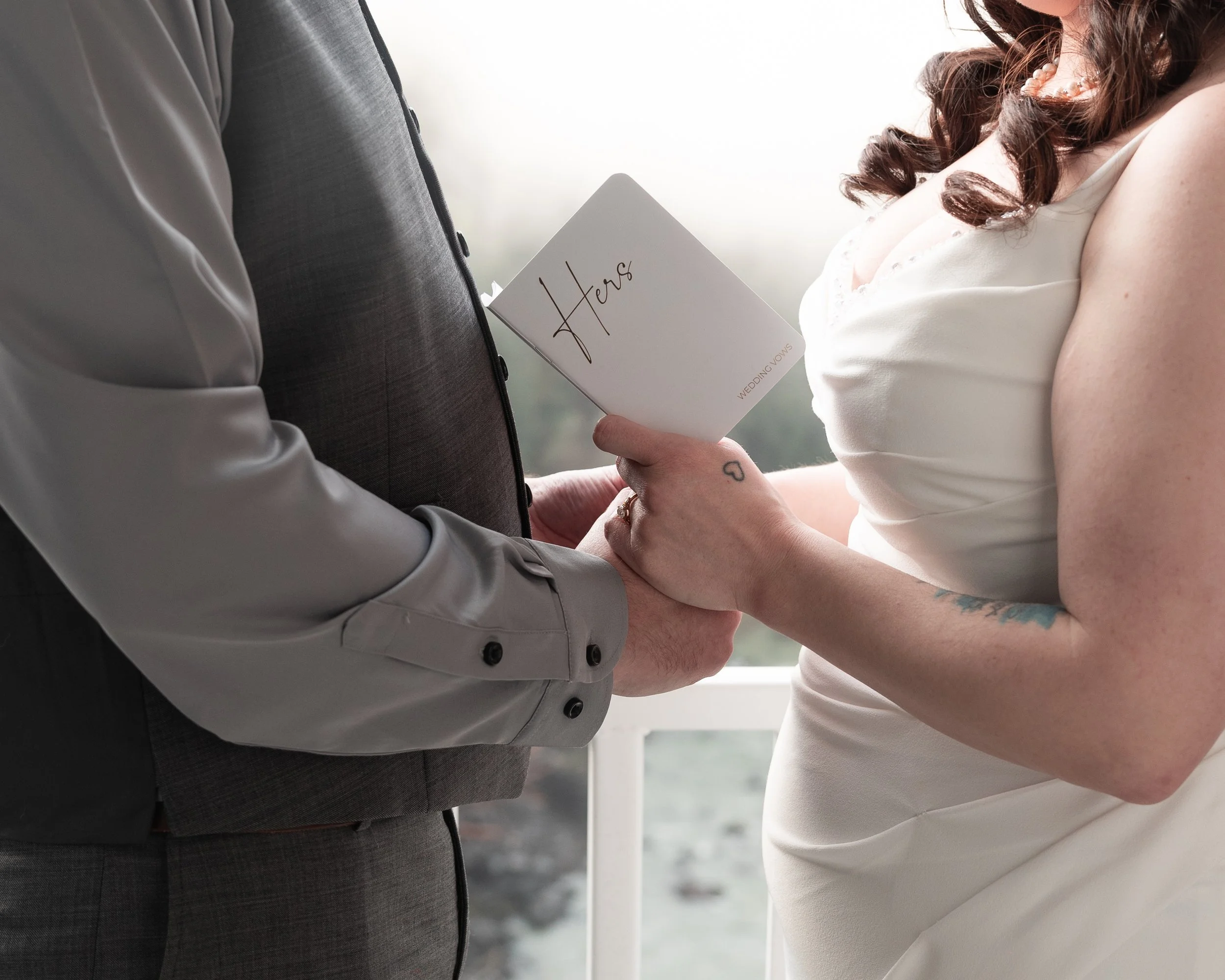 A bride and groom holding hands during their wedding ceremony, with the bride holding a booklet that says "Here" on the cover.