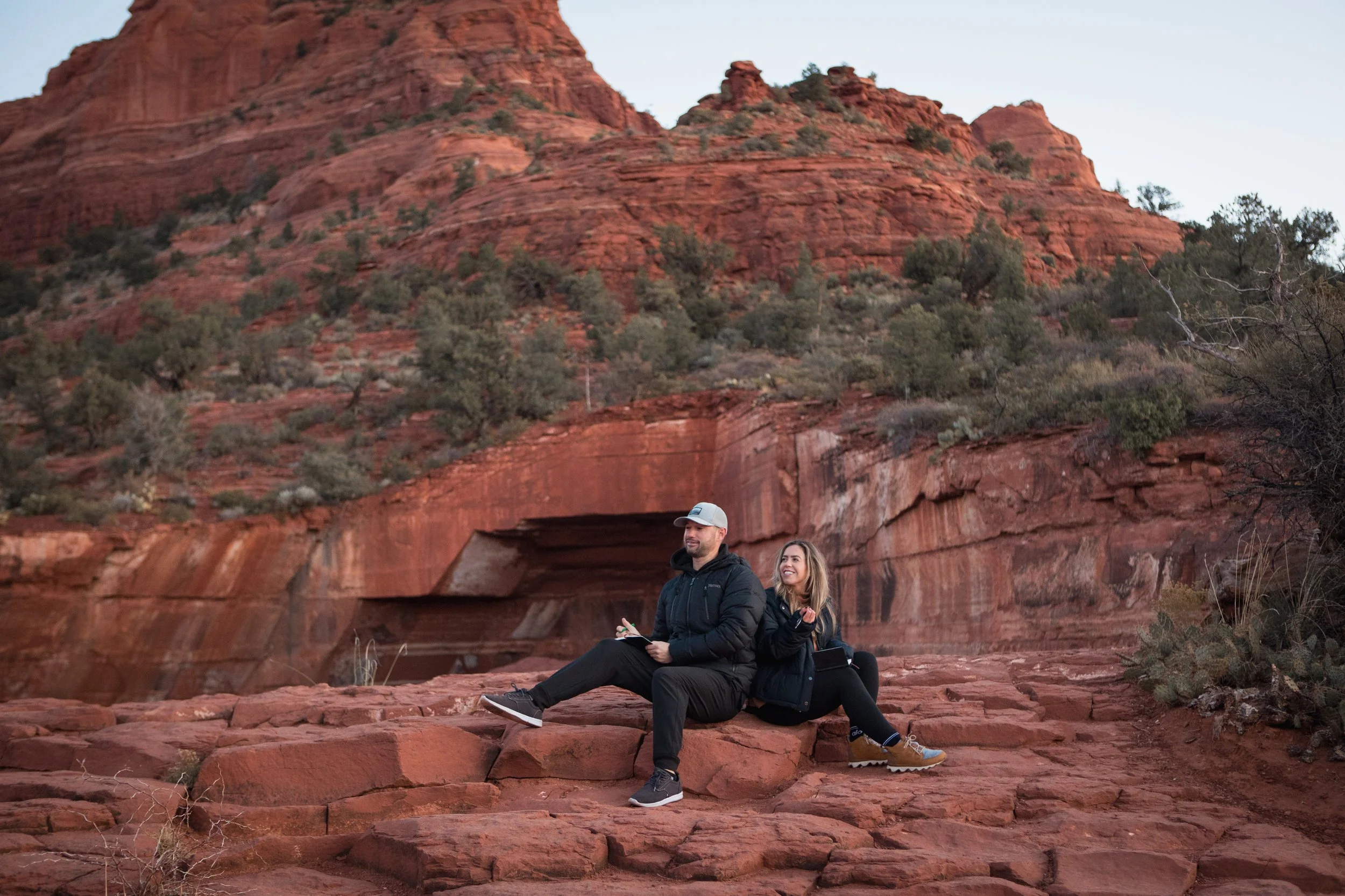 Couple writing their vows on a hike in the Sedona red rock, sitting back to back and smiling.