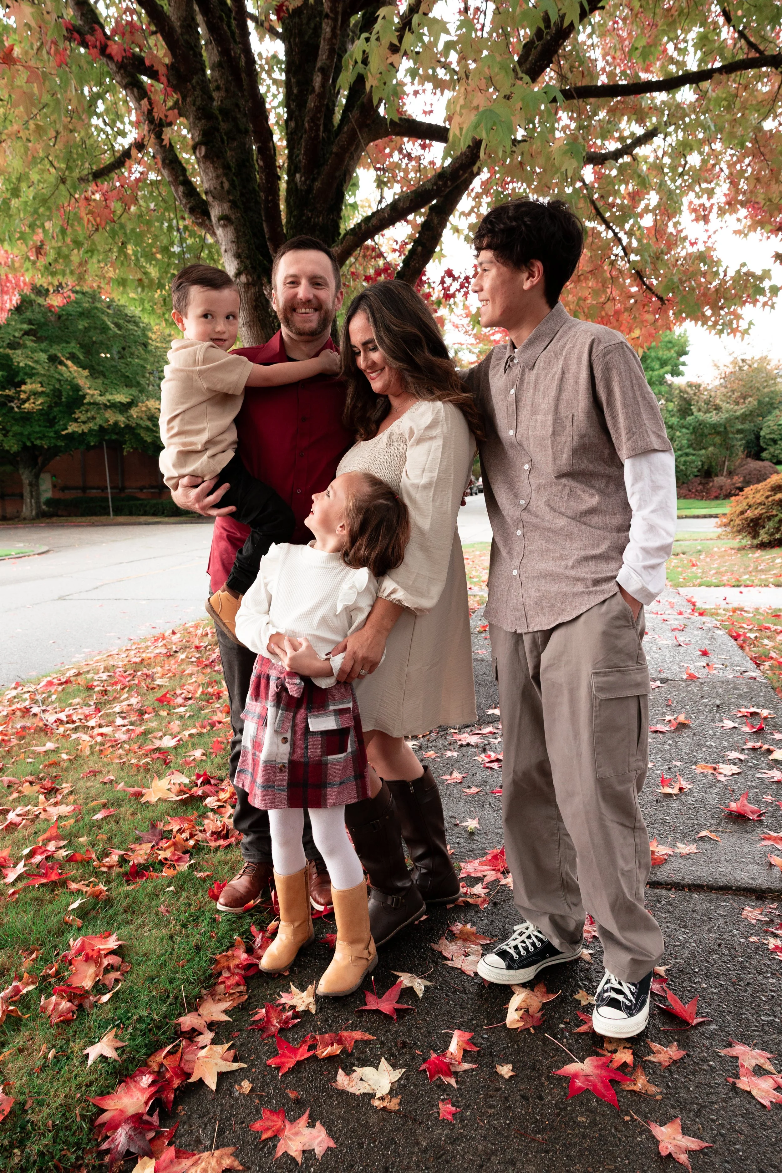 family of five in fall colors smiling at each other under the red and orange fall colored leaves of a maple tree
