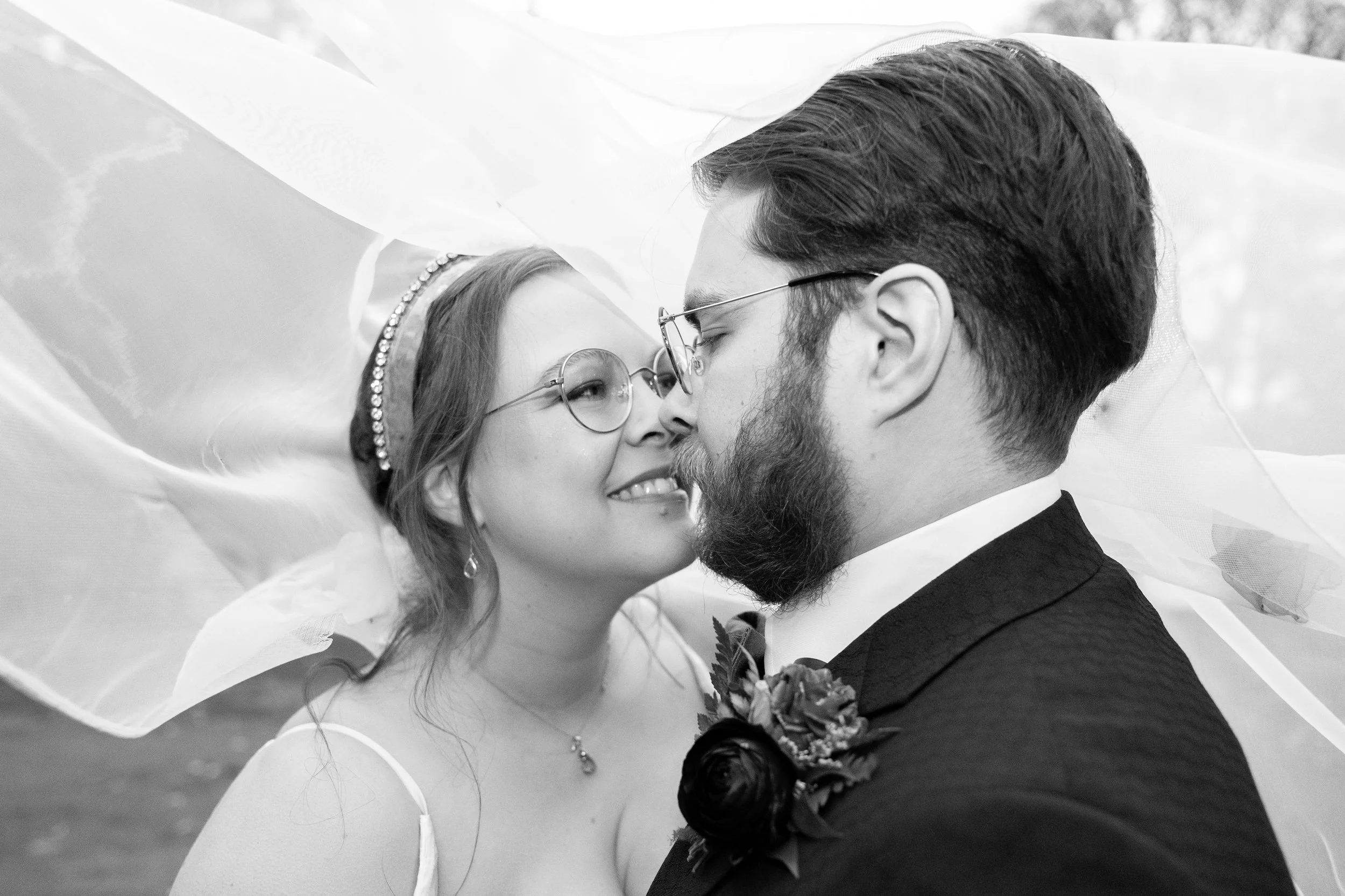 A black-and-white photo of a bride and groom close together, smiling and about to kiss, with a veil draped around them.