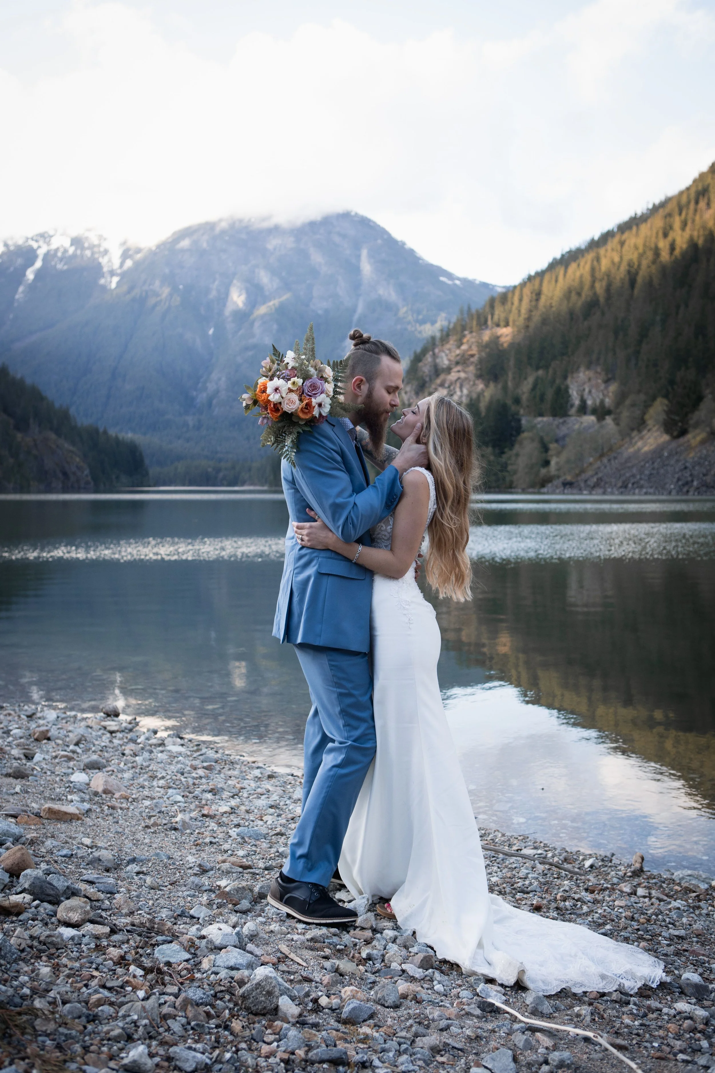 A bride and groom sharing a kiss near a mountain lake with mountains and forest in the background.