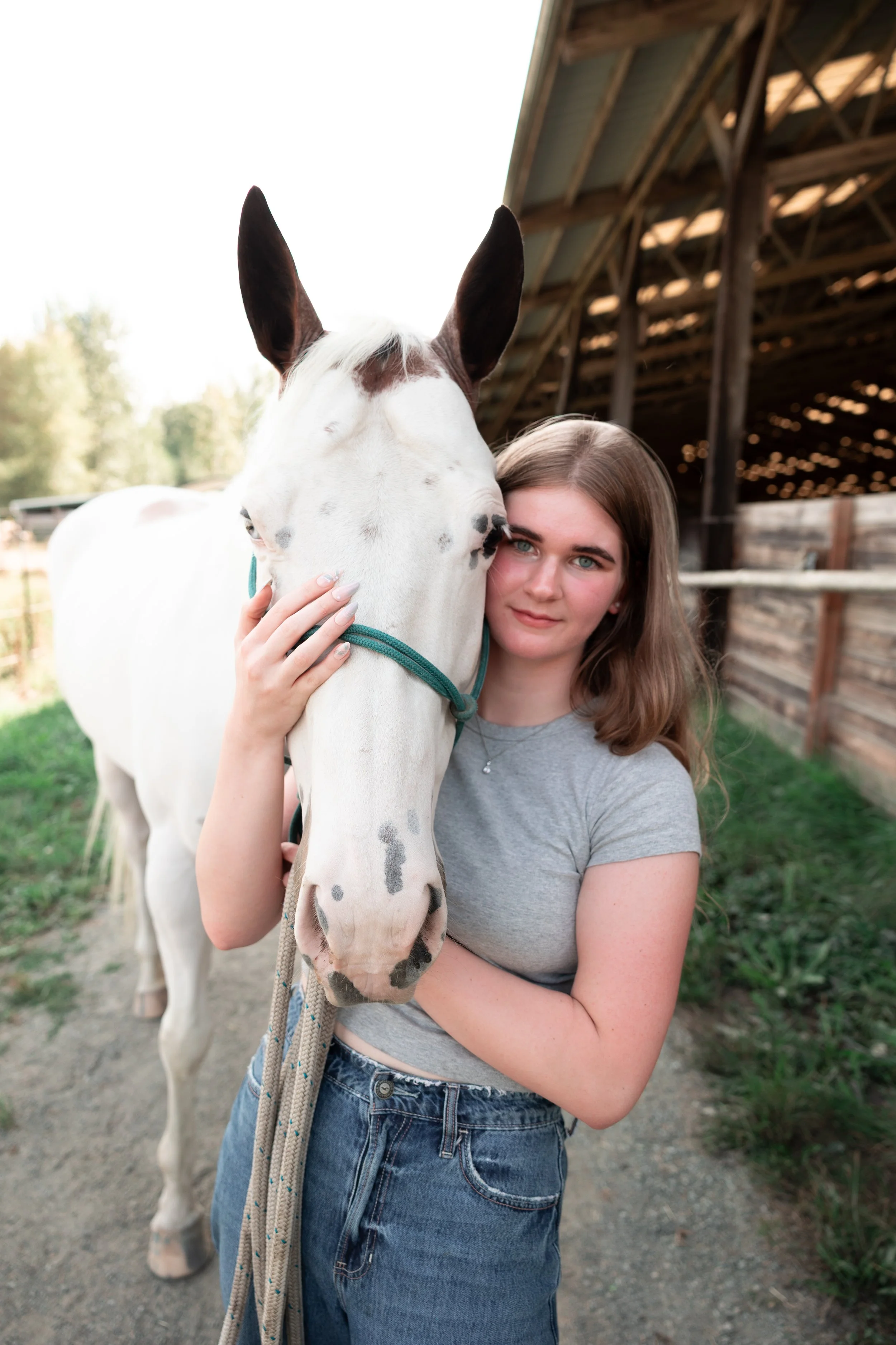 A young woman with long brown hair, wearing a gray t-shirt and jeans, is hugging a white horse with black spots, standing outdoors near a wooden barn. The woman is holding the horse's face gently, and they are both looking at the camera. The background shows green grass and trees.