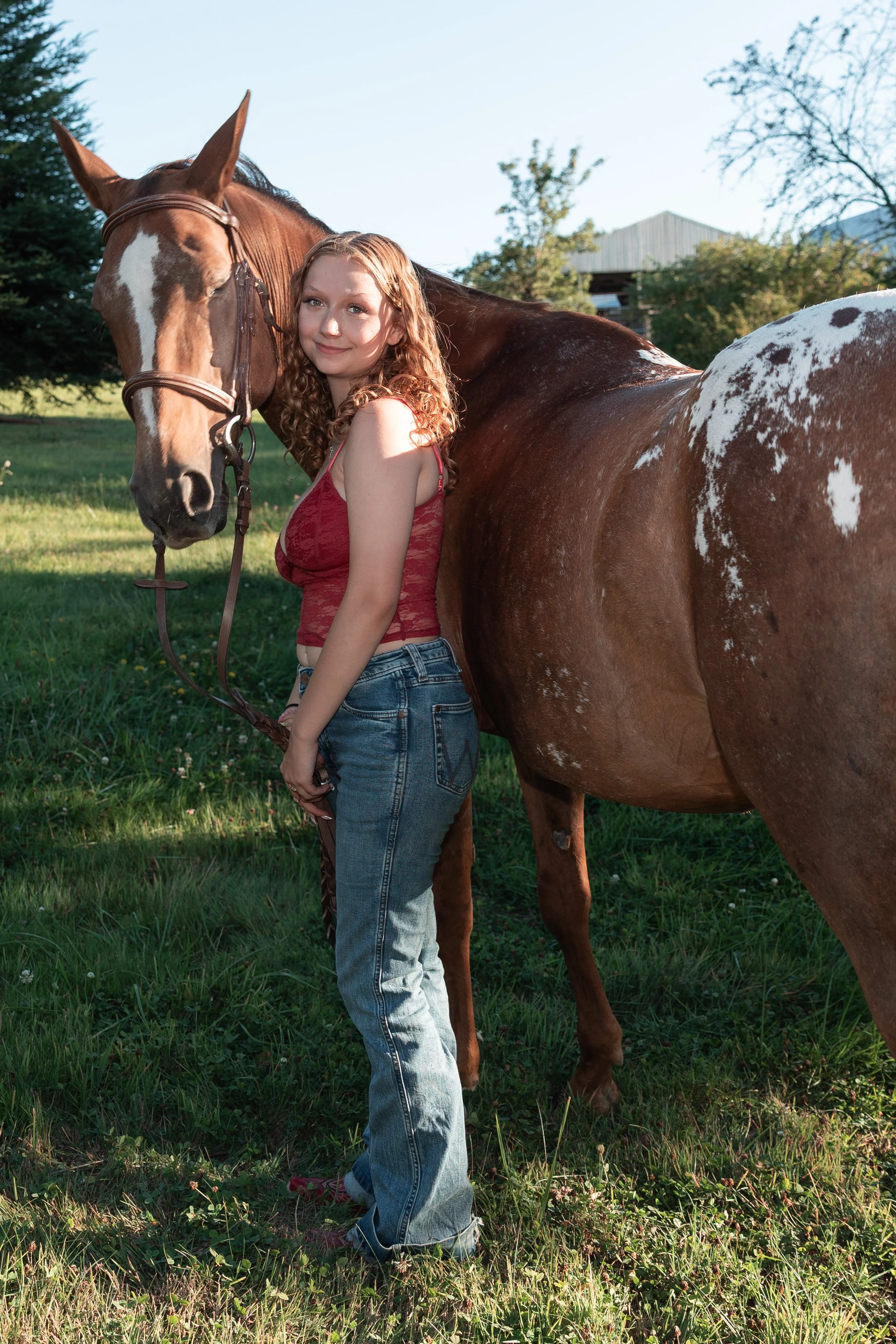 A young woman with curly red hair wearing a red crop top and jeans standing next to a brown and white pinto horse in a grassy field with trees and a barn in the background.