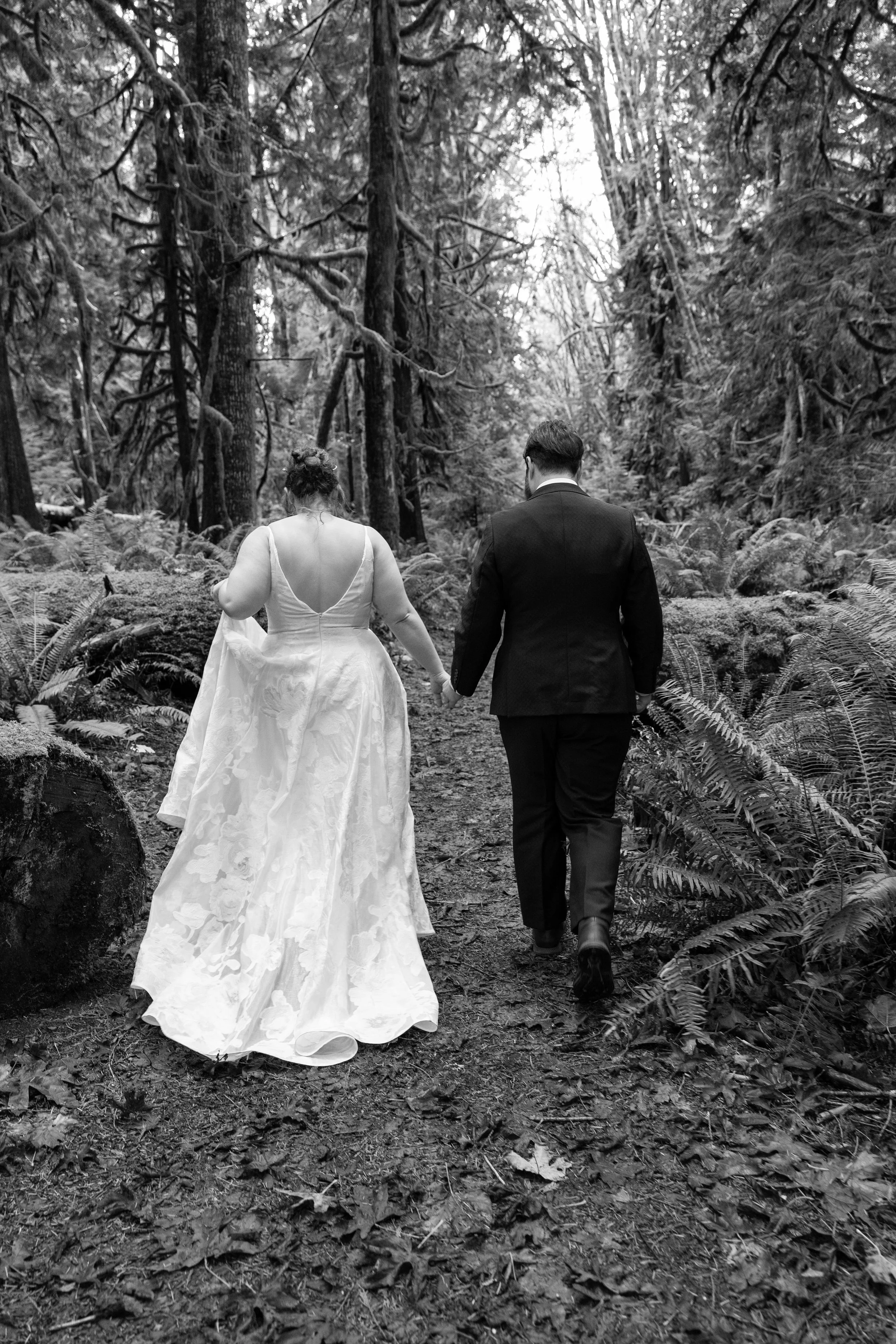 A couple, dressed in wedding attire, walking hand-in-hand down a forest trail.