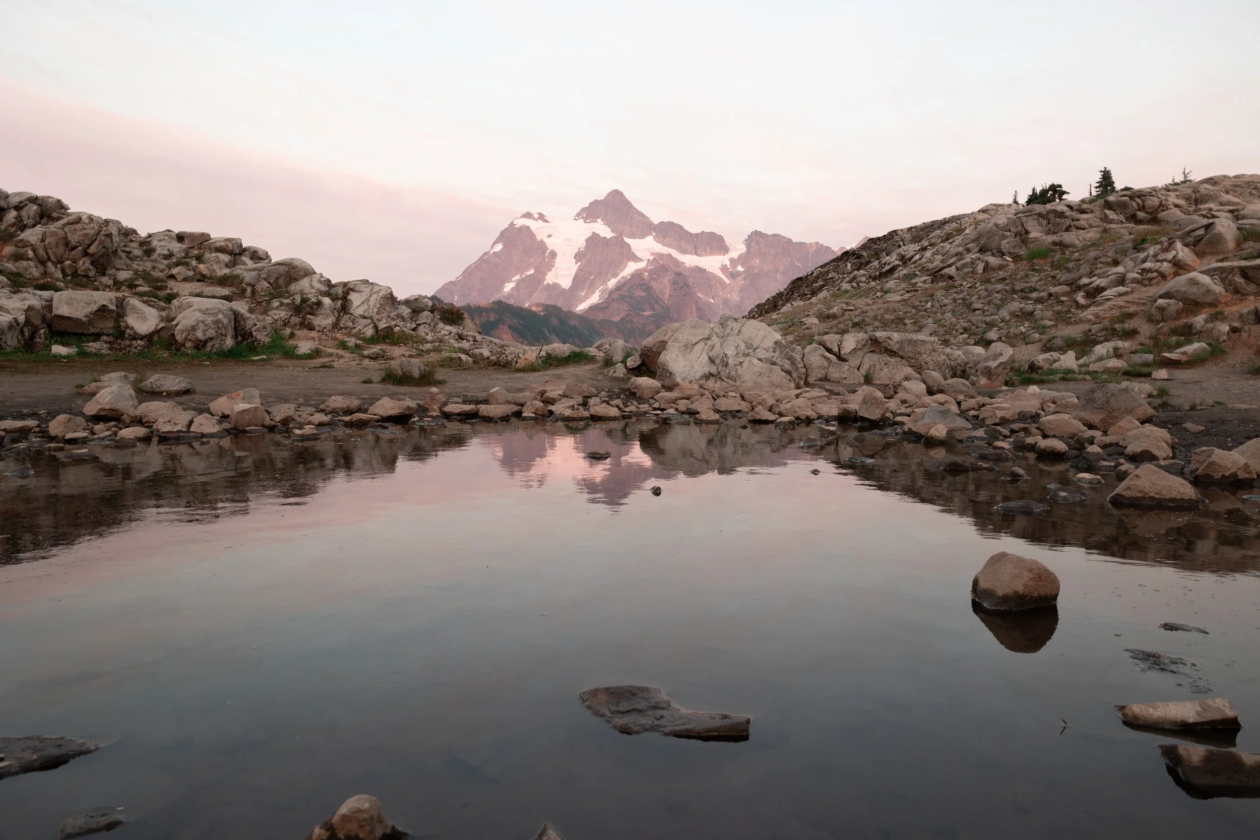 Scenic view at the top of Artist Point Mt.Baker WA with snow patches, rocky terrain, and a calm lake reflecting the sky and rocks in the foreground.