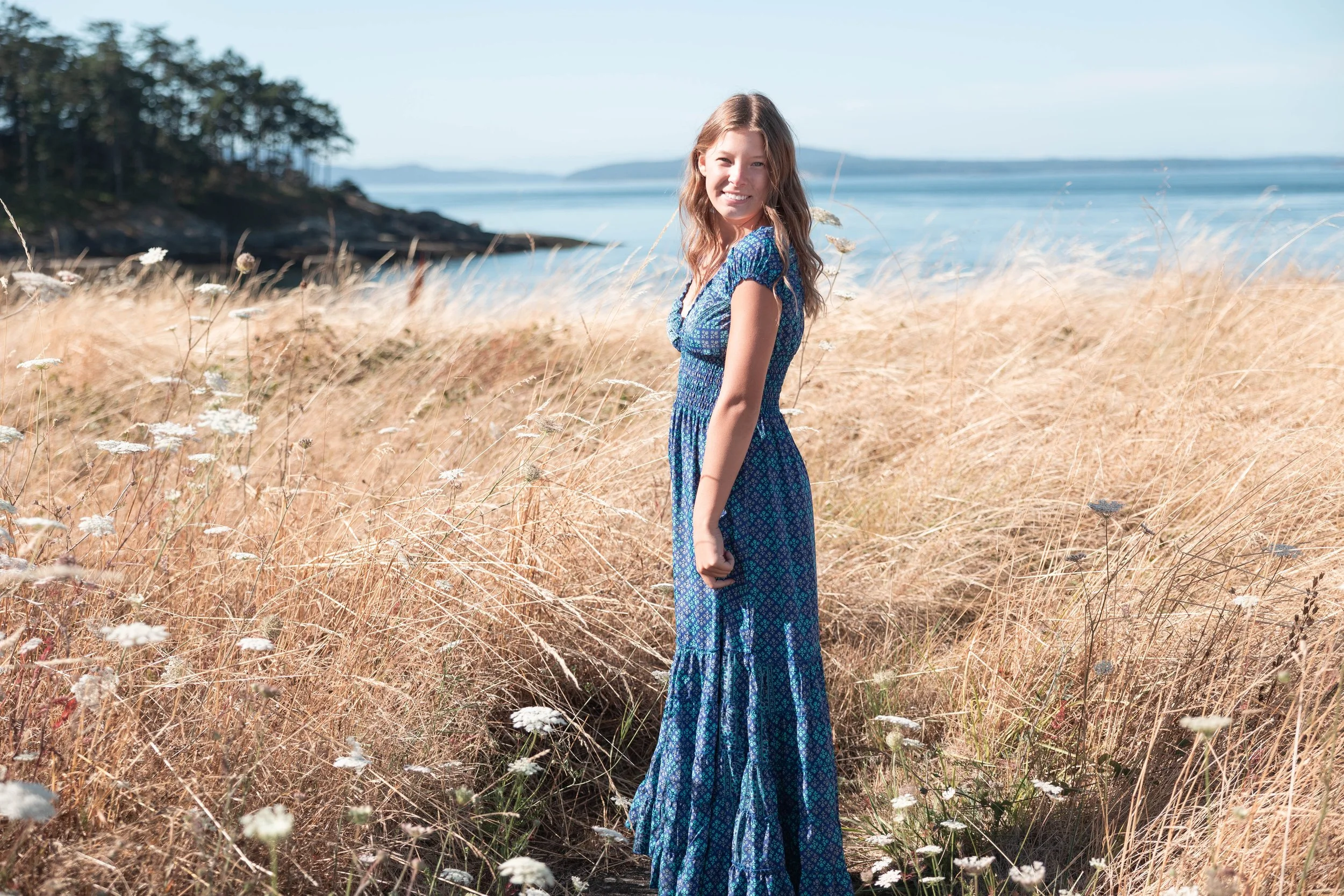High school senior girl in a long blue dress standing sidways smiling in a field with water and Orcas Island in the background