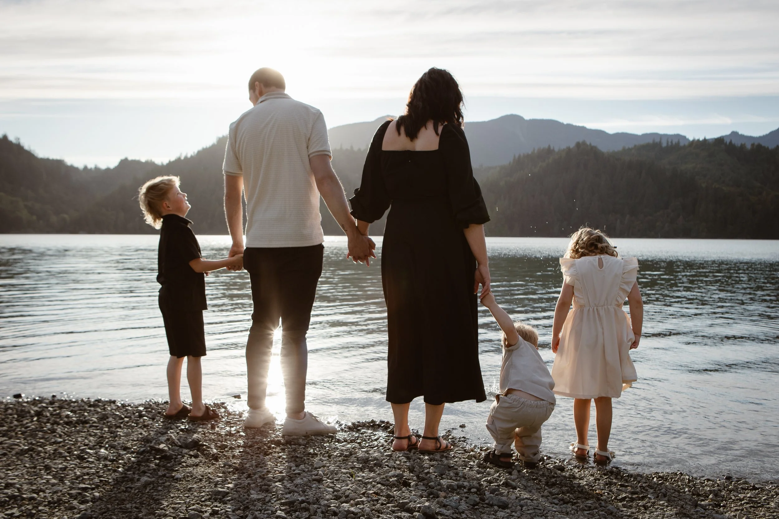 A family of six standing by a lake at sunset, holding hands and facing the water. The scene is scenic with mountains in the background and the sun low in the sky.