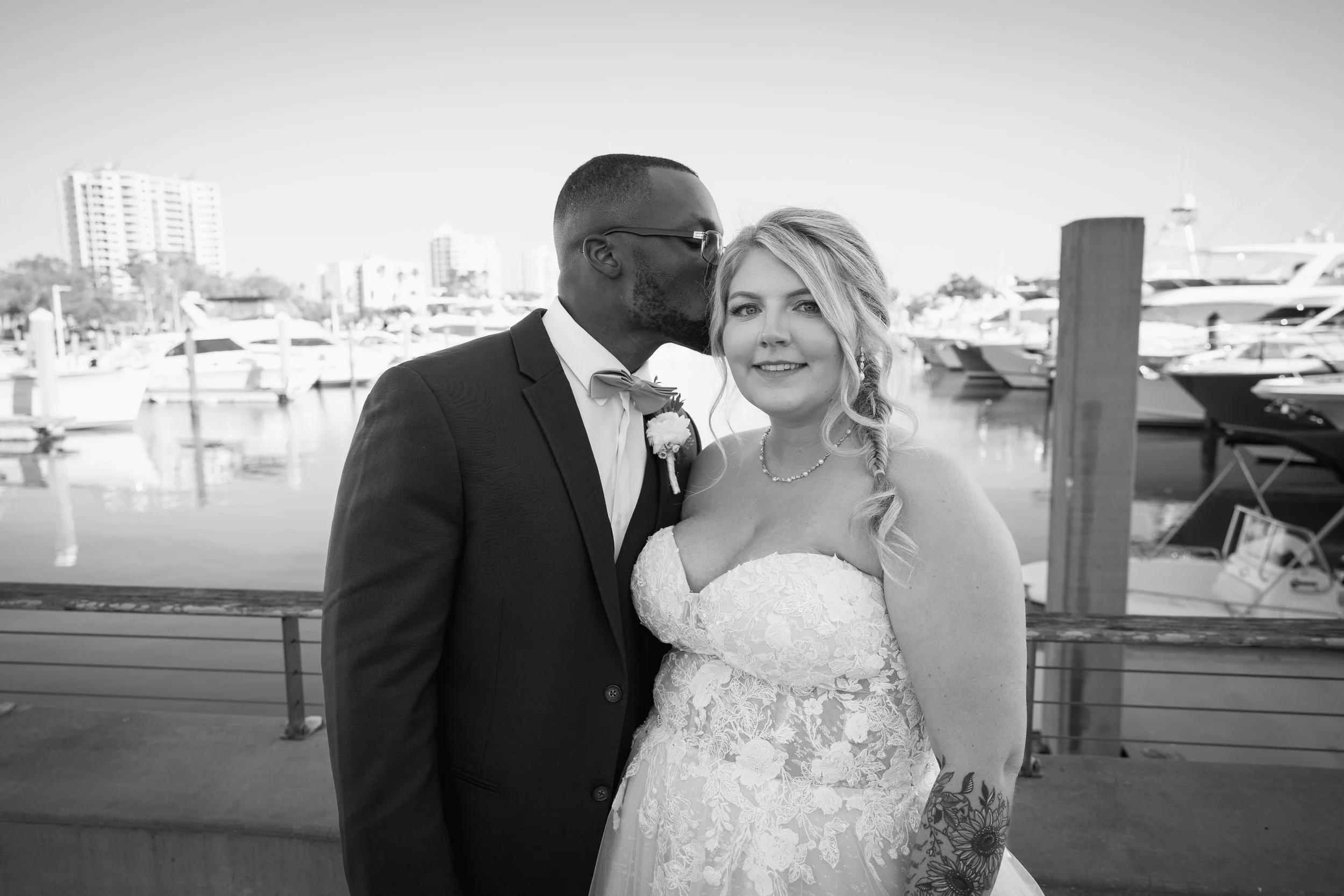 Black and white photo of a groom in a suit kissing a bride in a wedding dress at a marina with boats in the background