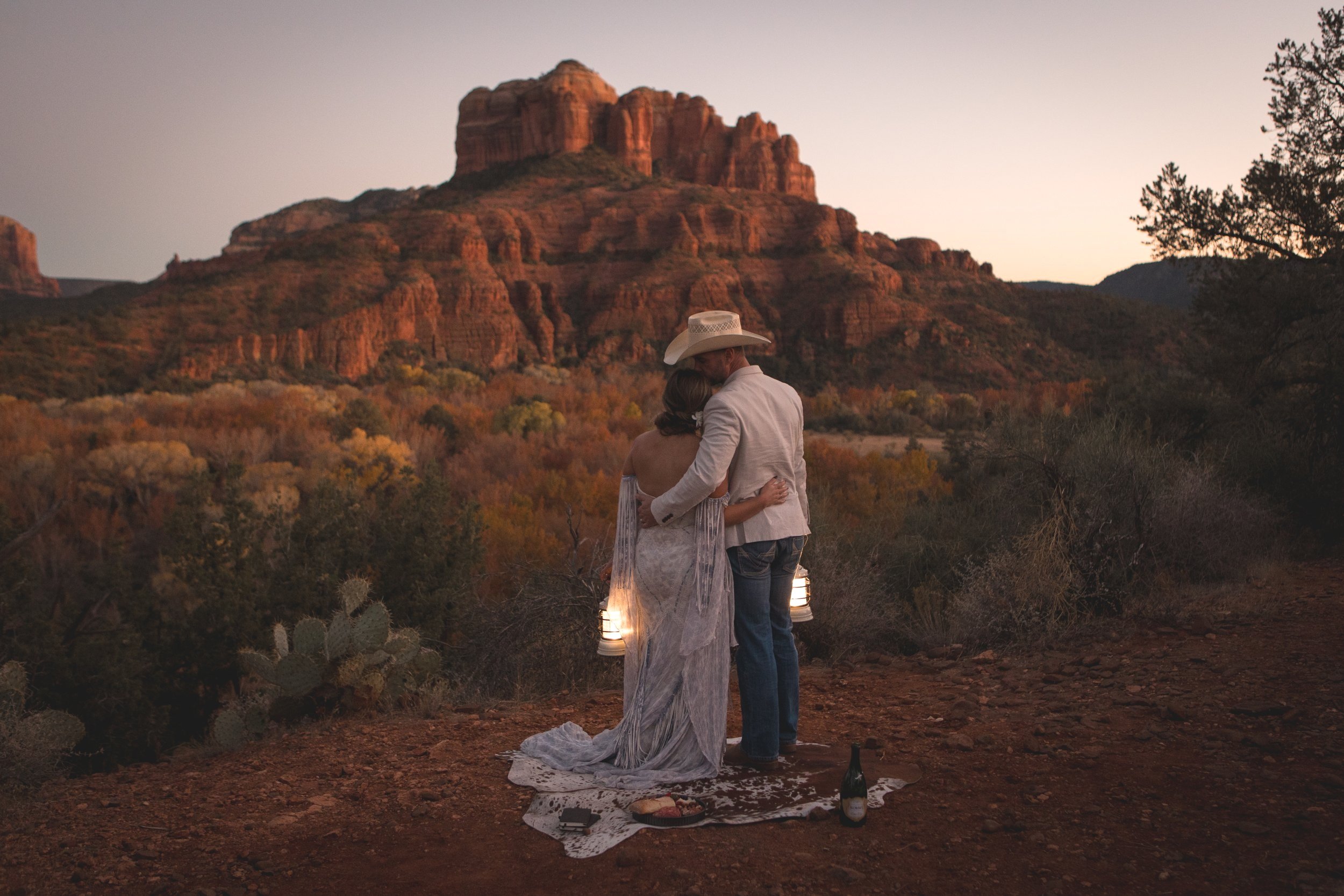 Sun setting on Sedon red rocks while the newly eloped couple holds each other with one hand and lanterns in the other while groom kisses her forehead
