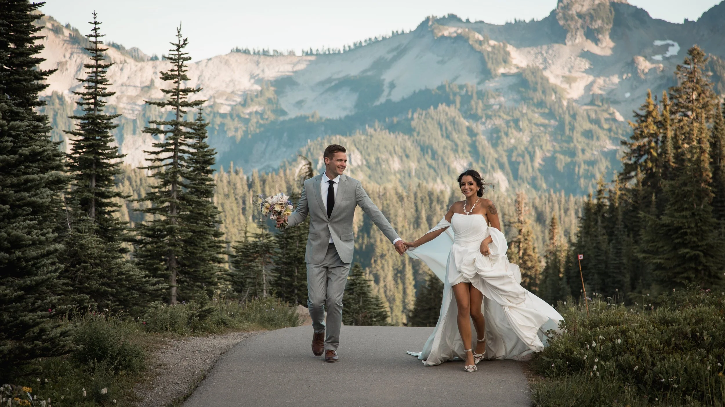 A newlywed couple walking hand-in-hand on a paved trail in a forested mountain area, with lush green trees and rocky mountain peaks in the background. The bride is wearing a white wedding dress and high heels, while the groom is dressed in a light gray suit with a black tie and brown shoes. The groom is holding a bouquet of flowers in his left hand, and they are smiling at each other.