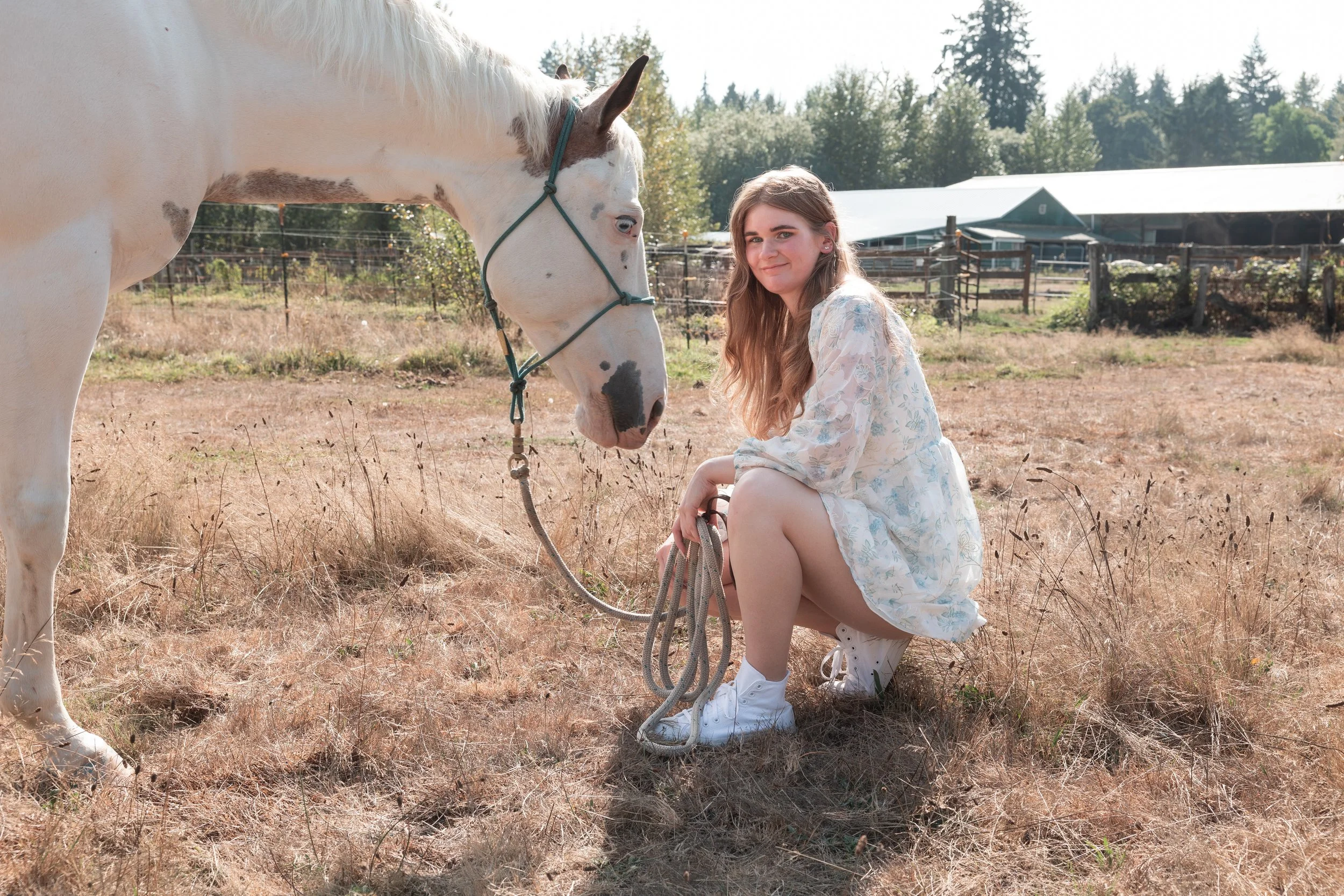 A young woman with long hair kneeling on one knee, holding the lead of a white horse with brown spots on a farm or ranch. The horse is facing the woman and they appear to be interacting. In the background, there are trees, a metal building, and a wooden fence, with a clear daylight sky.