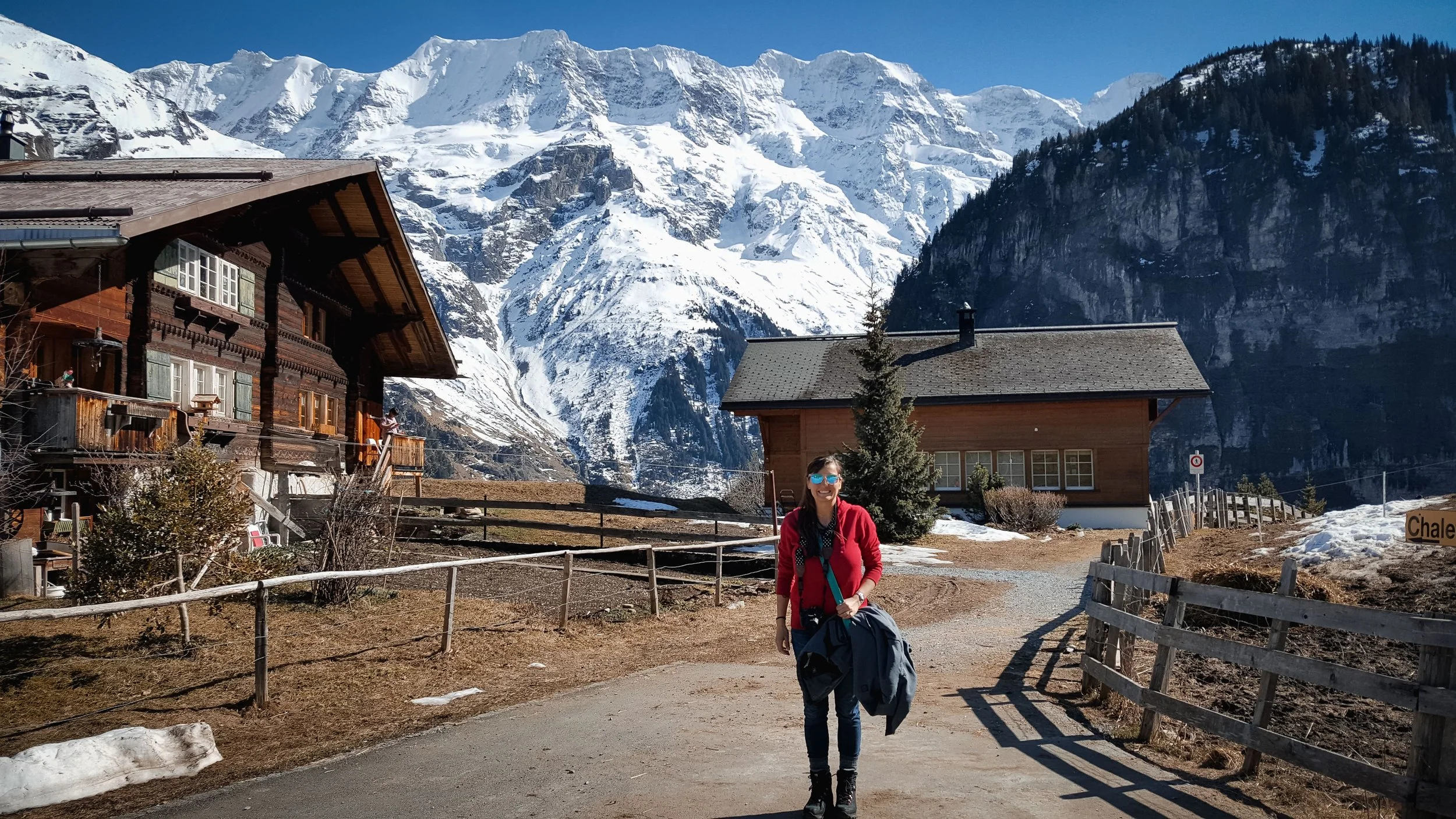 Bernadette Micro-wedding photographer, wearing sunglasses and a red jacket stands outdoors in the Swiss Alps in Switzerland,with snow-capped peaks, wooden chalet houses in background.
