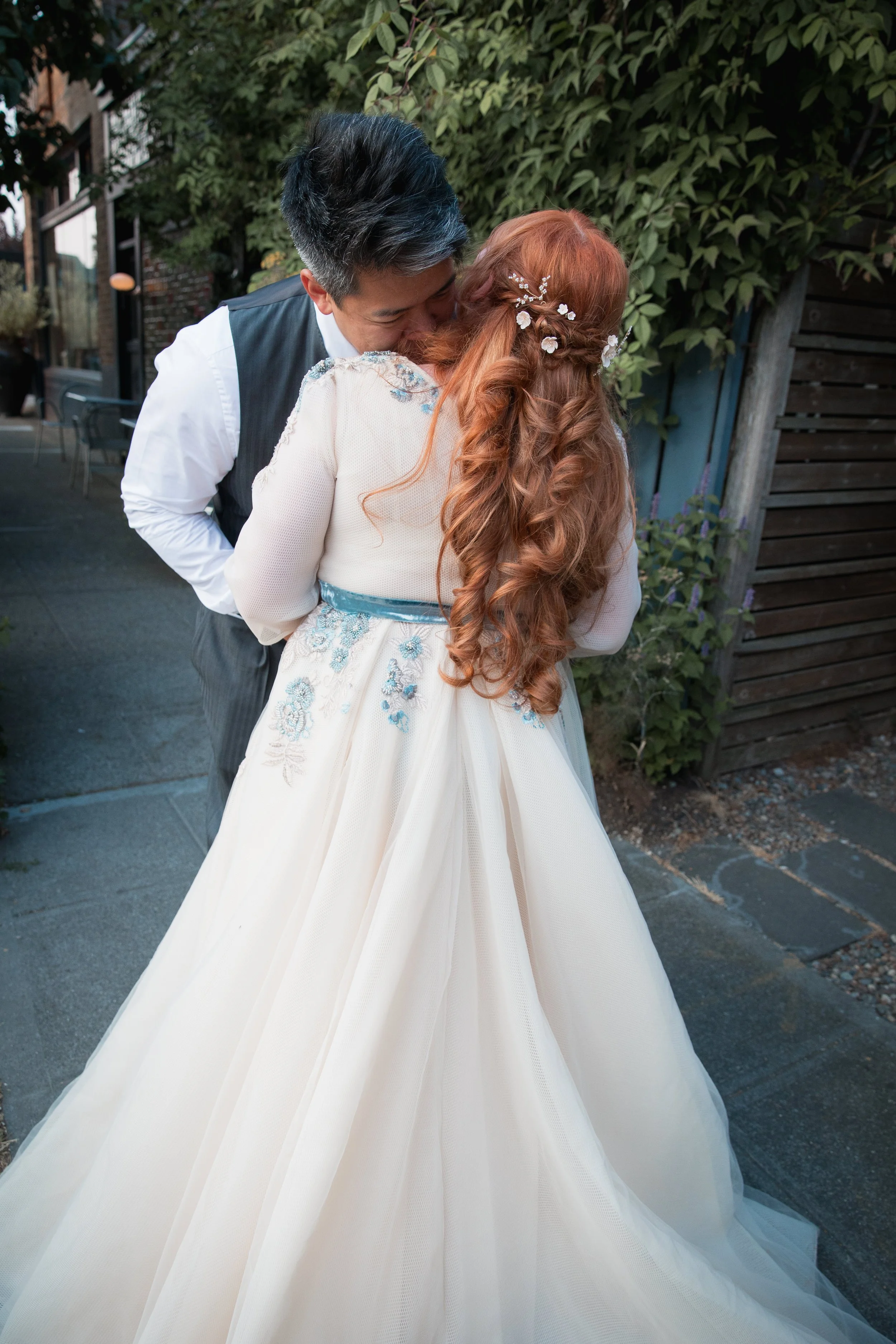 A couple sharing an intimate moment outdoors, with the woman in a white wedding dress with blue embroidery, and the man in a white shirt and dark vest, surrounded by greenery.