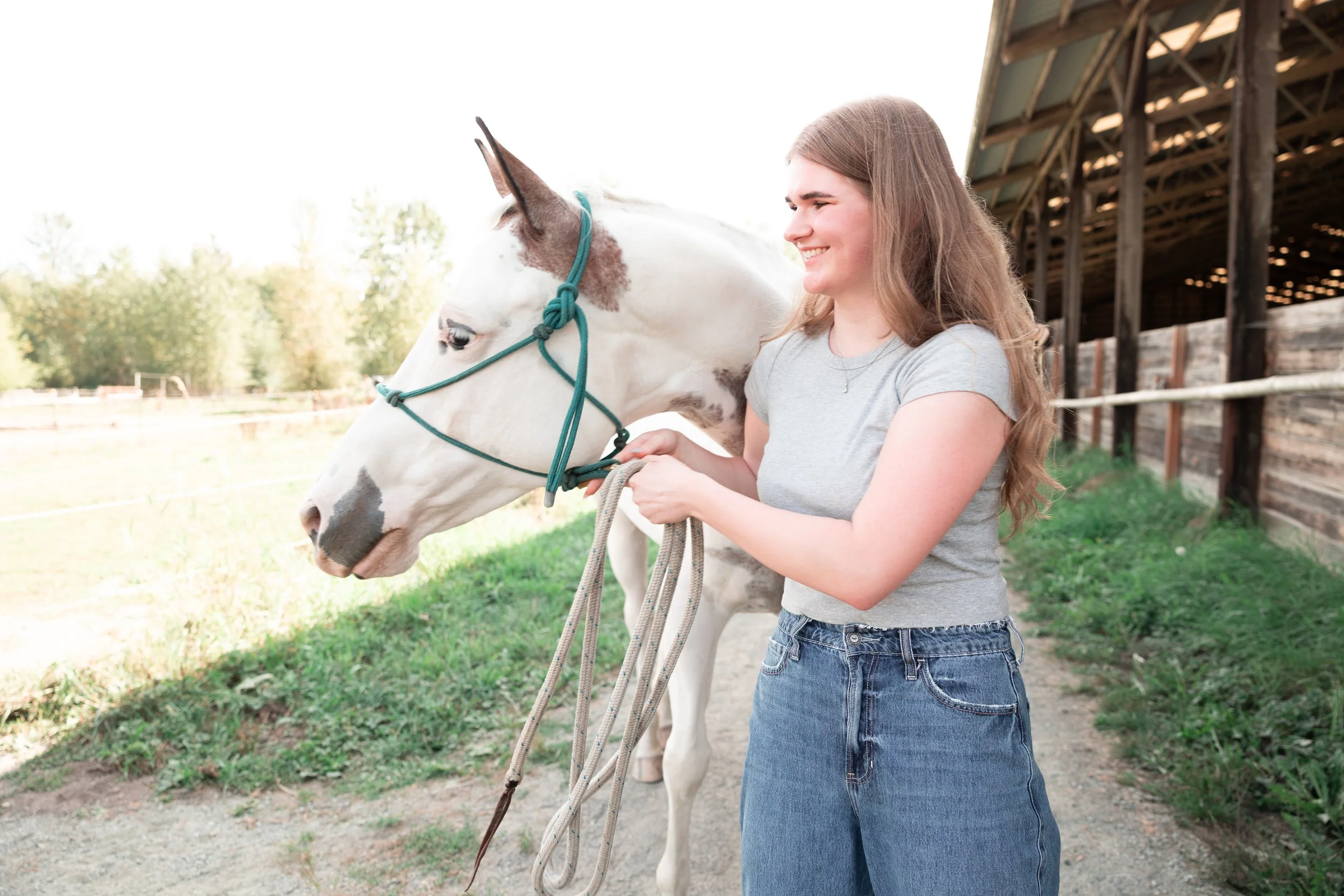 A young woman with long hair smiling and holding the reins of a white and brown horse in a farm setting with a barn in the background.