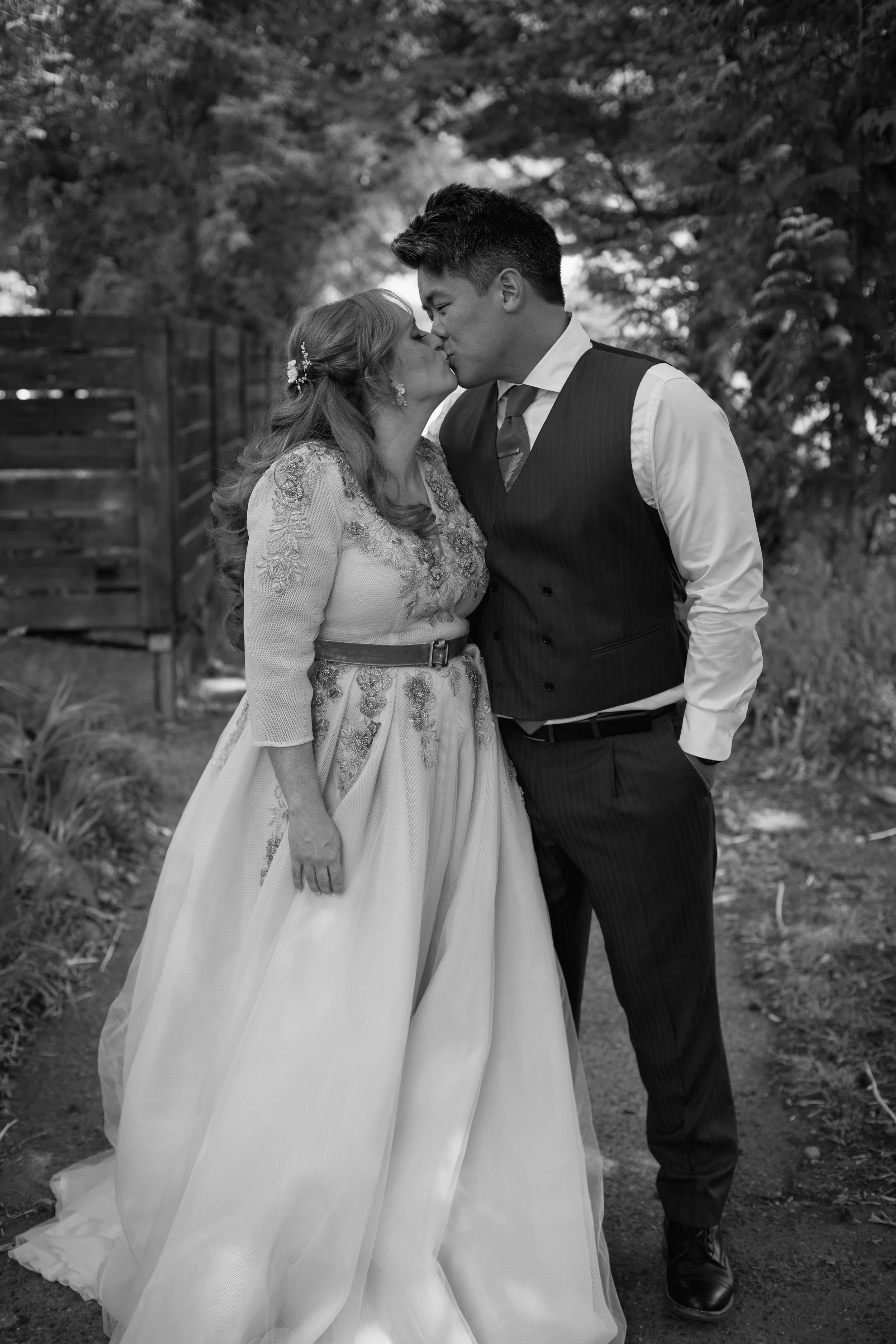 A couple dressed in wedding attire sharing a kiss outdoors.