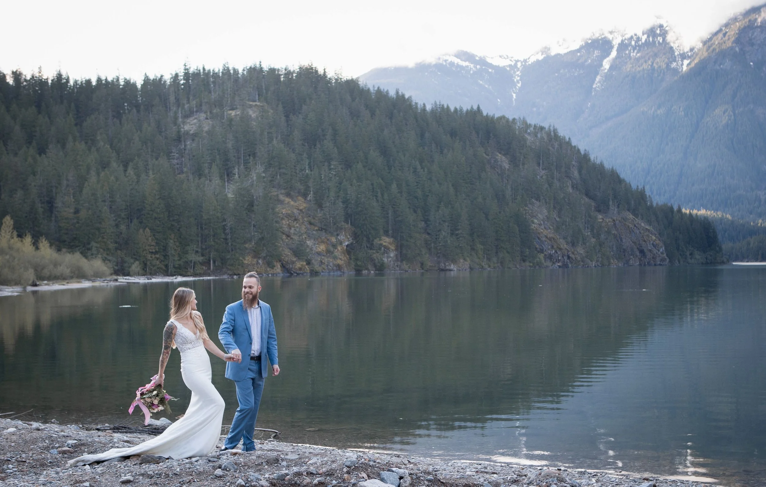 A newlywed couple holding hands on a rocky lakeshore with a forested mountain landscape and snow-capped peaks in the background.