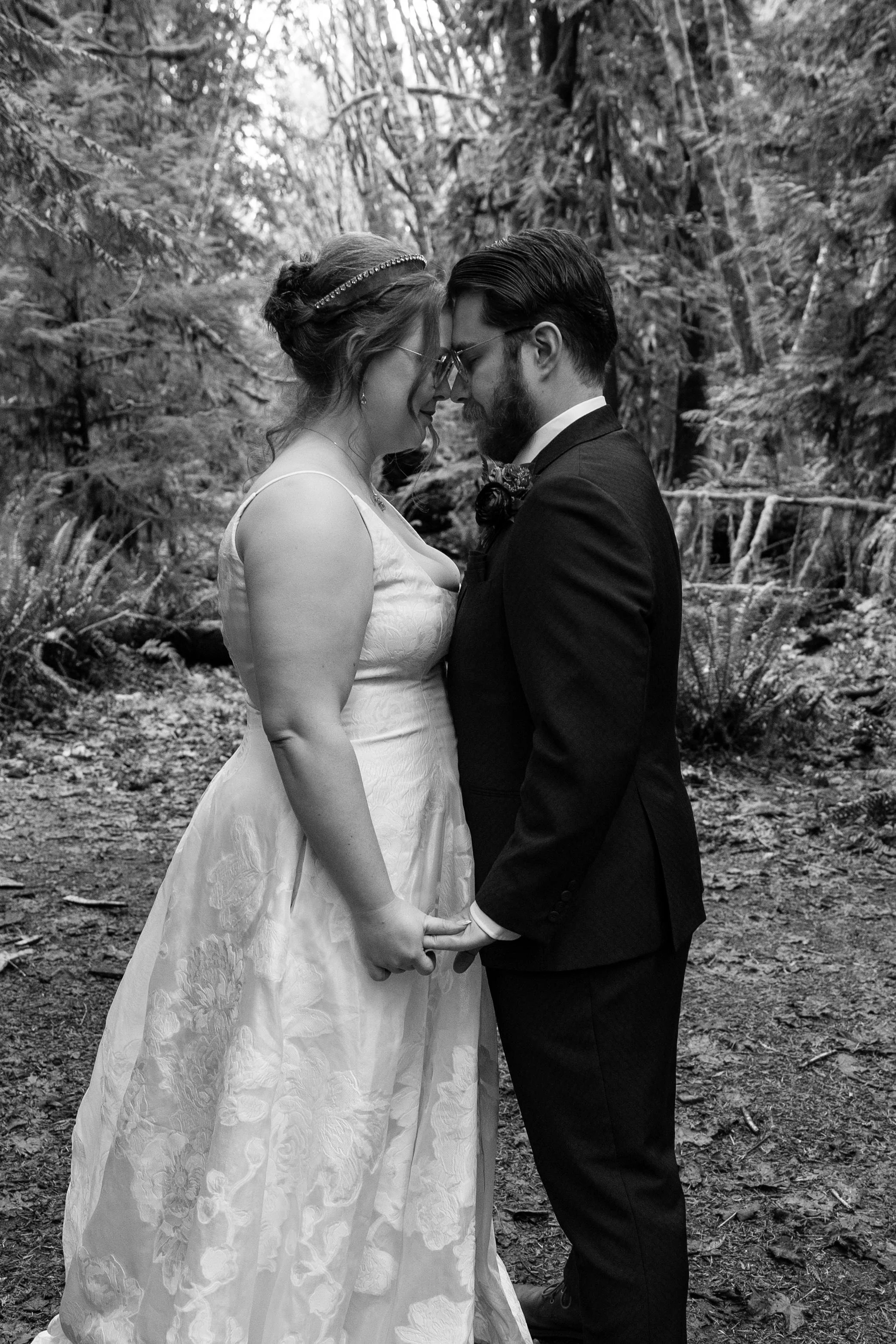 Black and white photo of a newlywed couple standing close together in a forest, holding hands with foreheads touching.