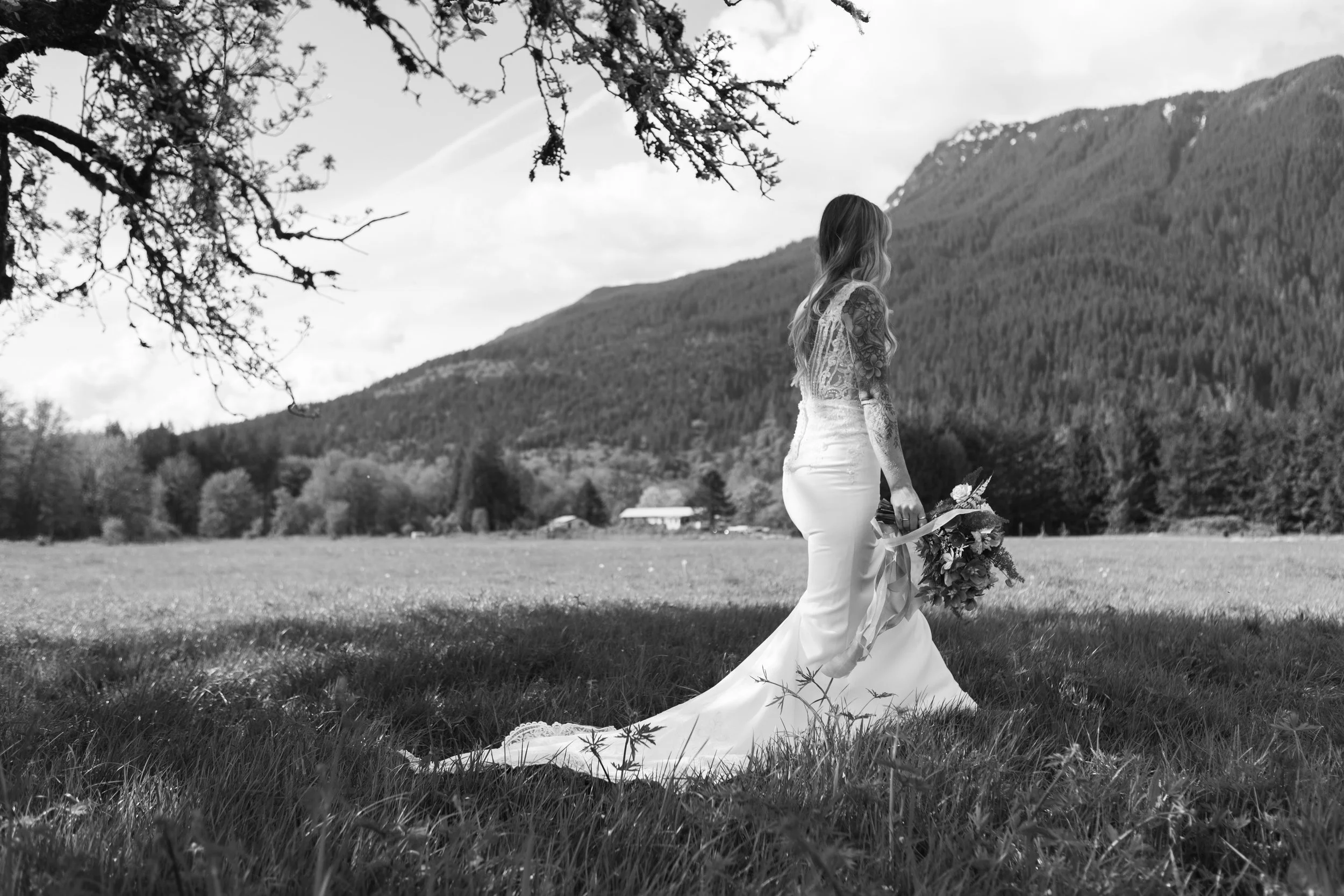 A woman in a wedding dress holding a bouquet, standing alone in a grassy field near mountains in black and white.