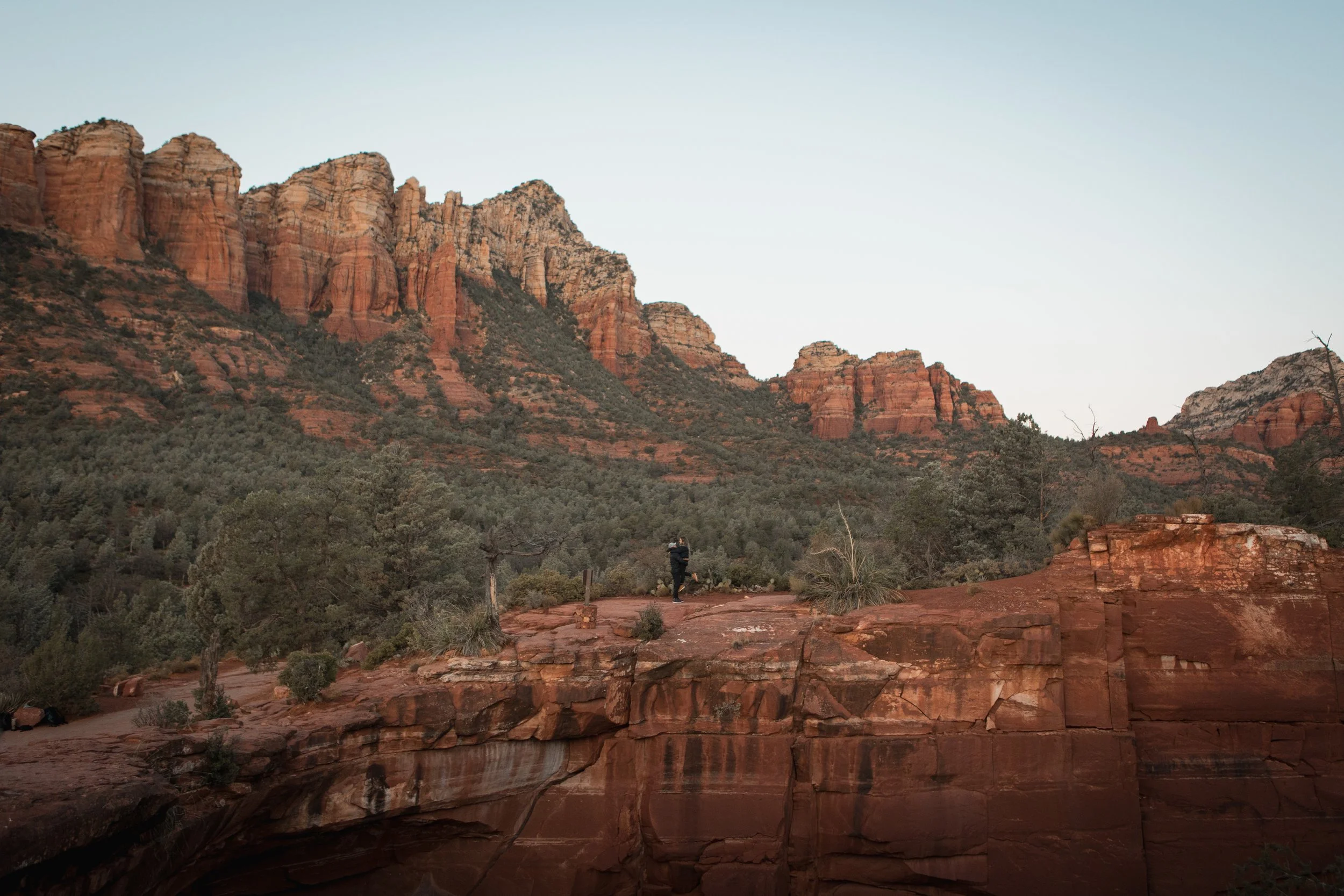 Sedona red rock landscape with a couple in the distance, he is picking her up and her legs are lifted behind her. They are in black hiking clothes.