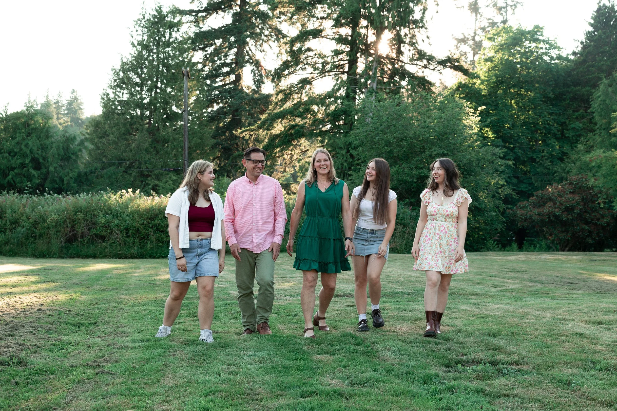 family of 5 waking towards camera looking at each other and laughing while the sun is setting behind the trees in the background