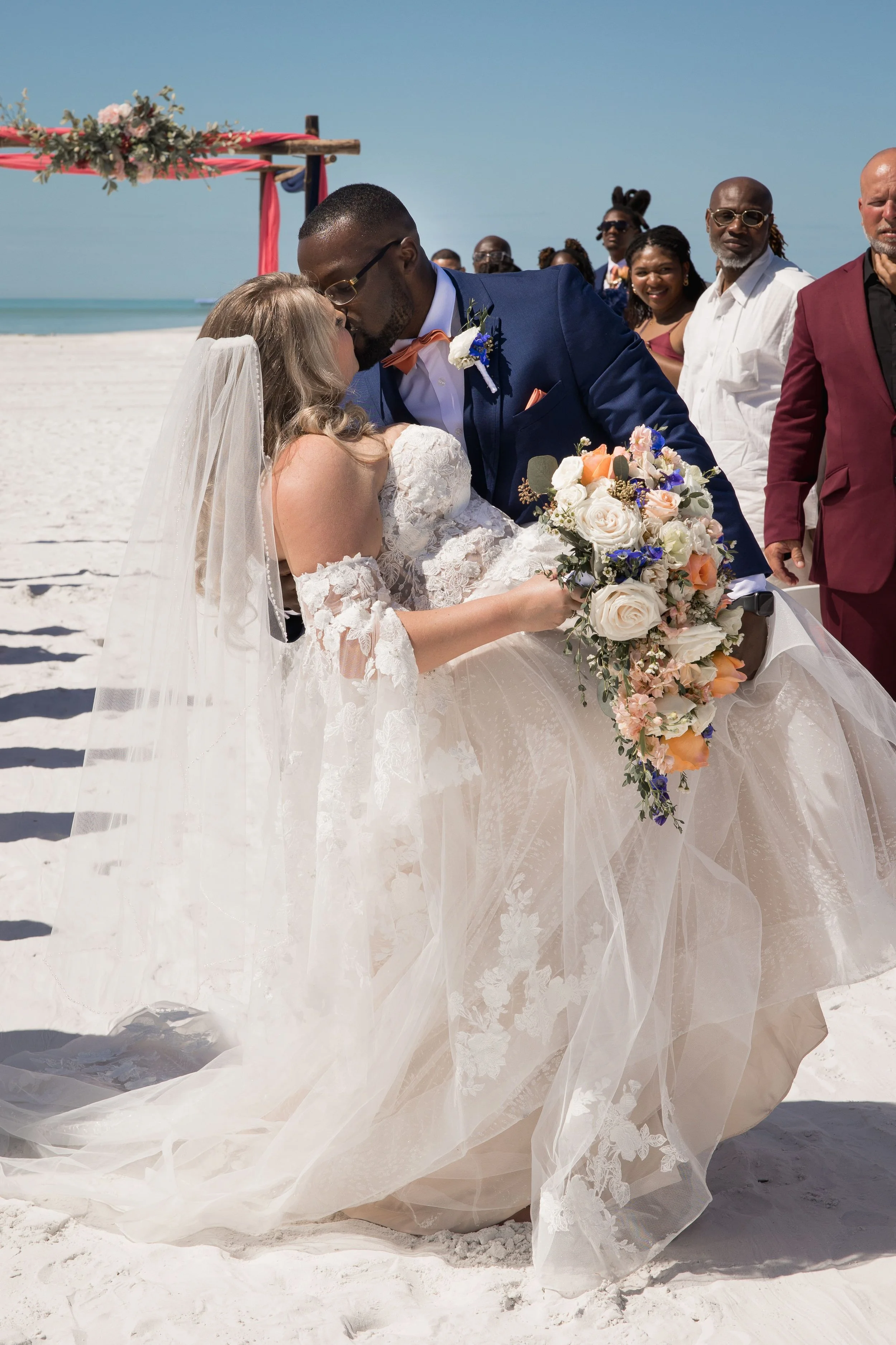 A bride and groom kissing during their beach wedding ceremony, with guests in the background on a sunny day.