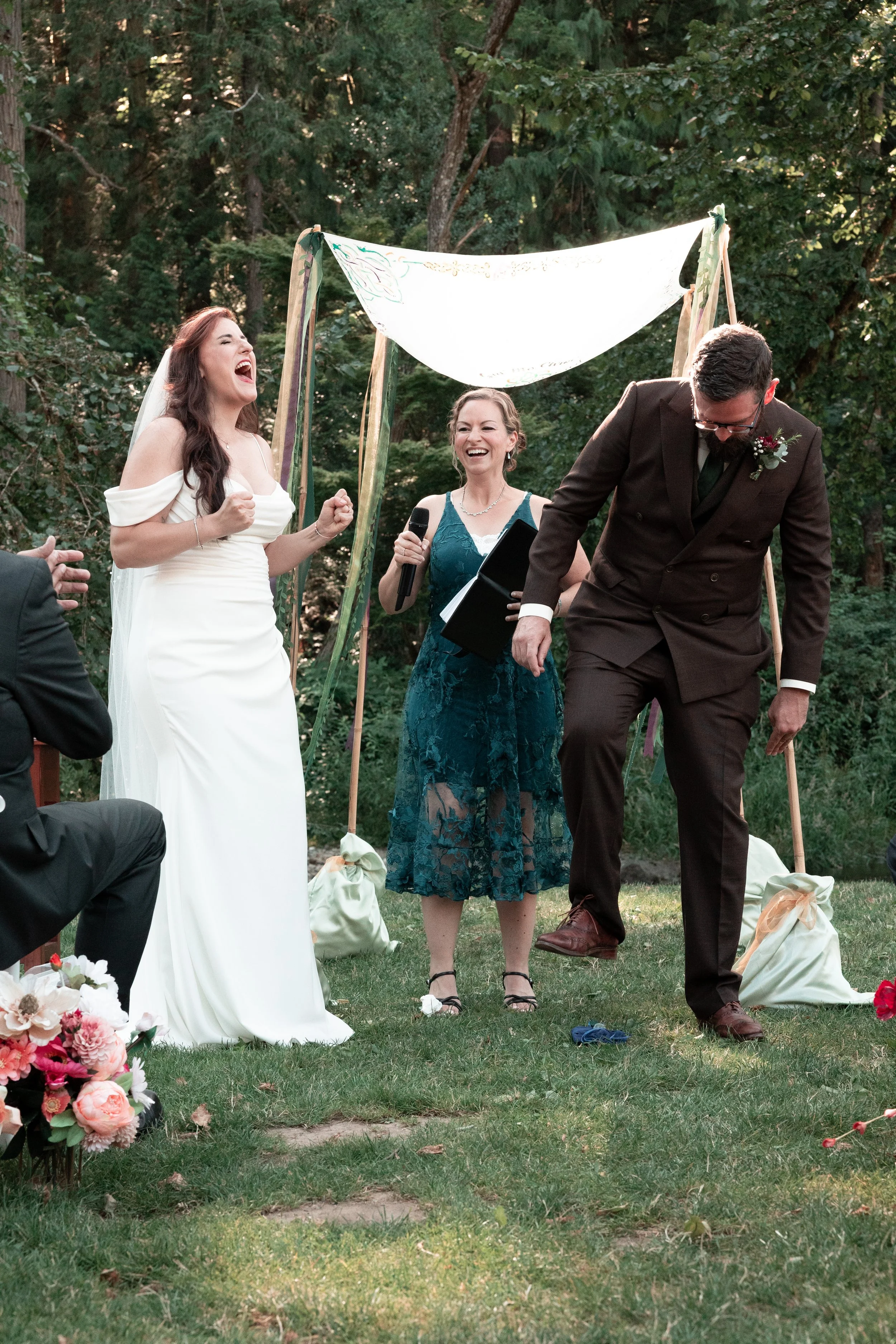 Bride and groom at an outdoor wedding ceremony, with a woman officiant or speaker in the middle, under a small decorative arch, surrounded by greenery and flowers.