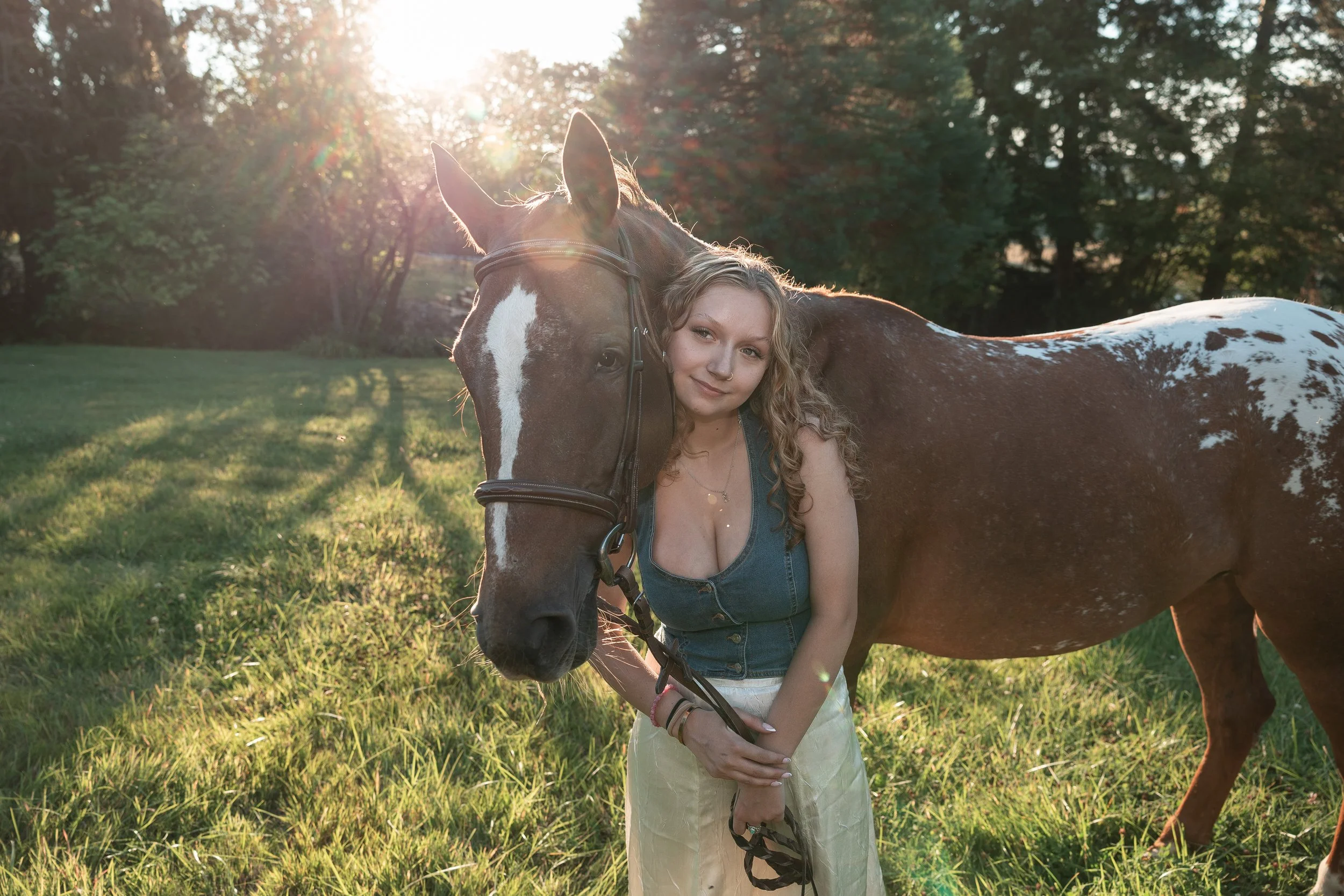 A young woman with curly blonde hair and a nose ring stands close to a brown and white spotted horse in a green field. The woman is smiling faintly, wearing a denim vest and yellow pants. The sun is setting or rising in the background, casting a warm glow and creating lens flare through the trees behind them.
