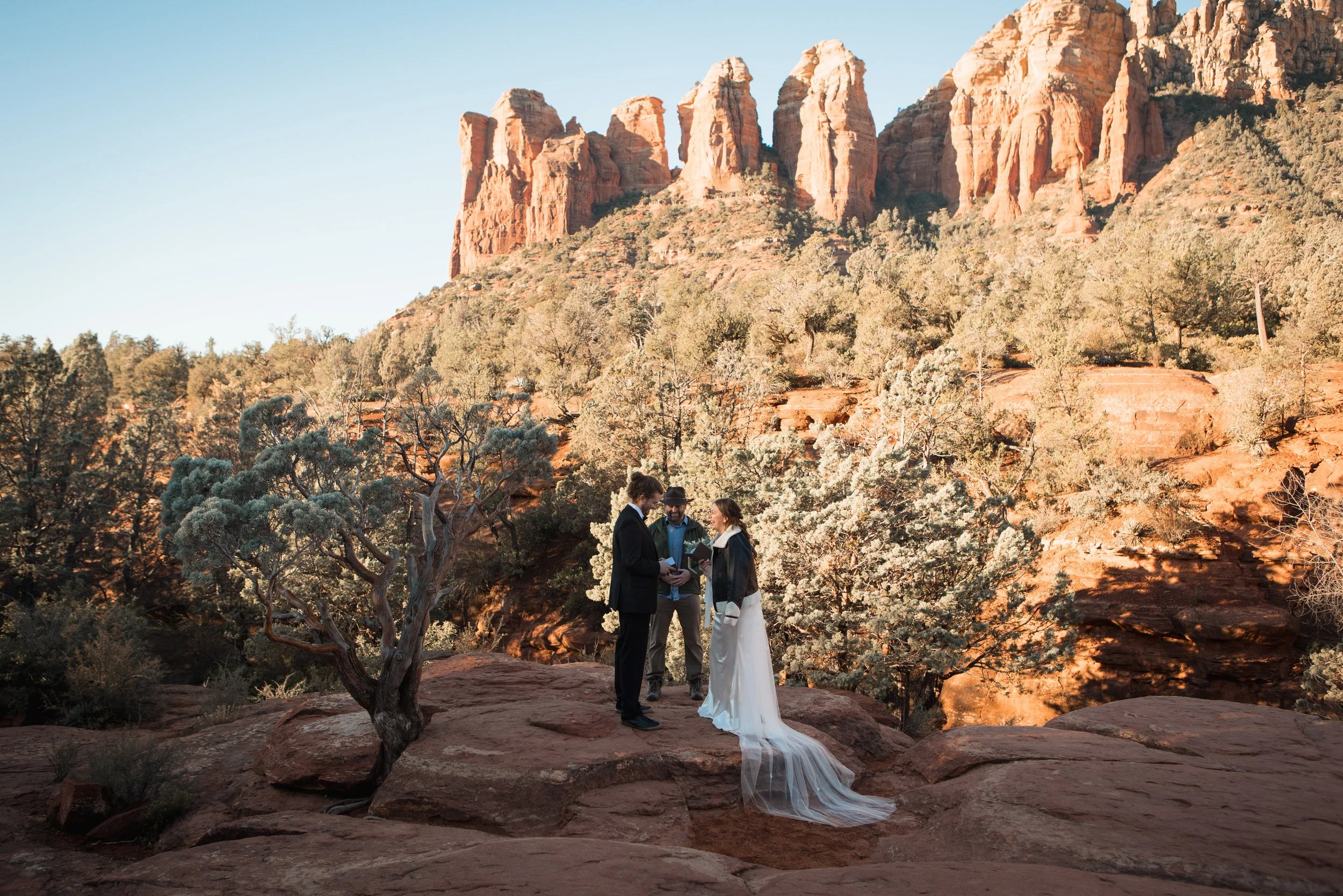 A Sedona couple in wedding attire exchanging vows in a desert landscape with red rocks and trees. An officiant stands nearby, with towering red rock formations in the background.