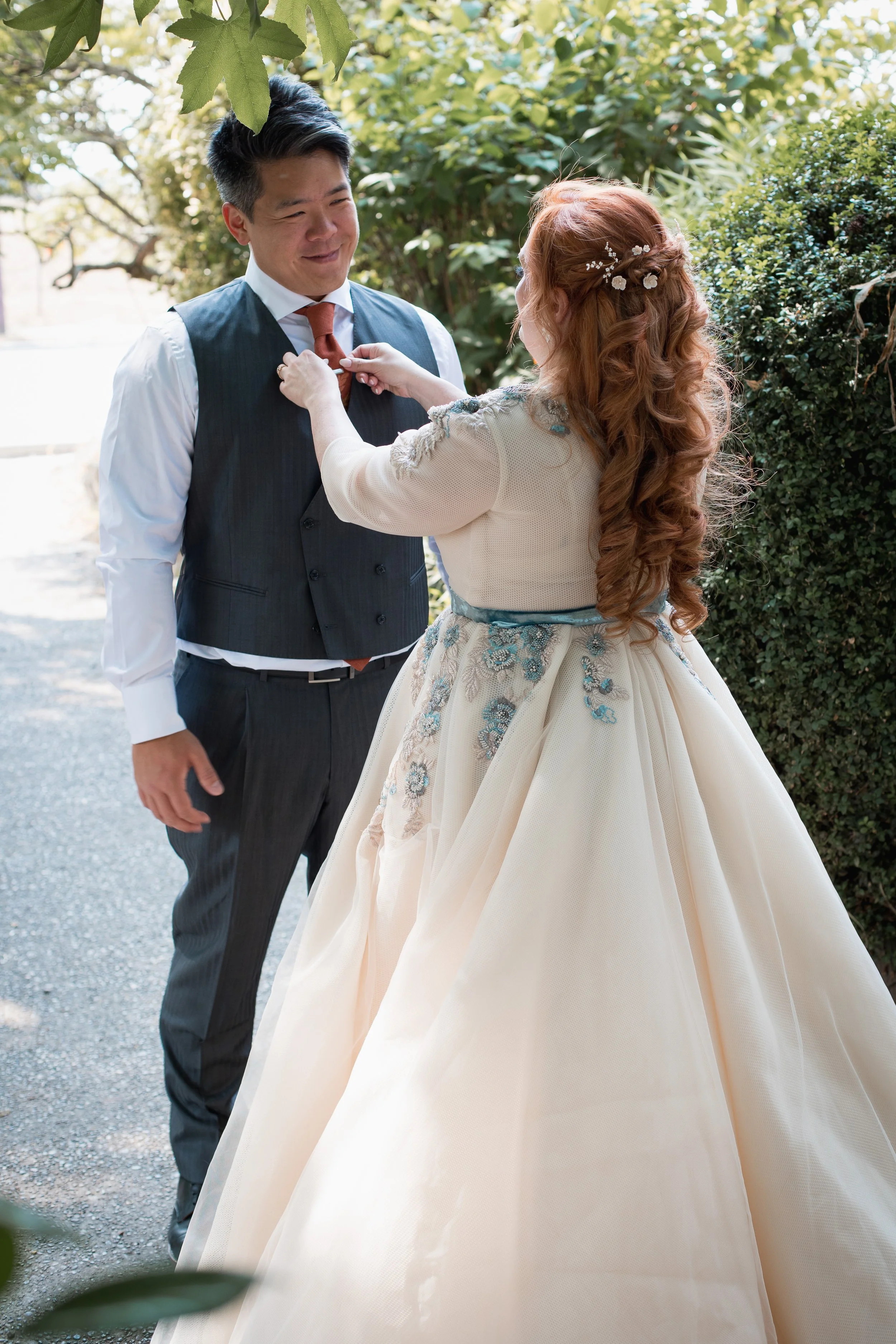 A bride in a white wedding dress with blue floral embroidery adjusting the groom's tie outdoors while standing on a gravel path surrounded by green foliage.