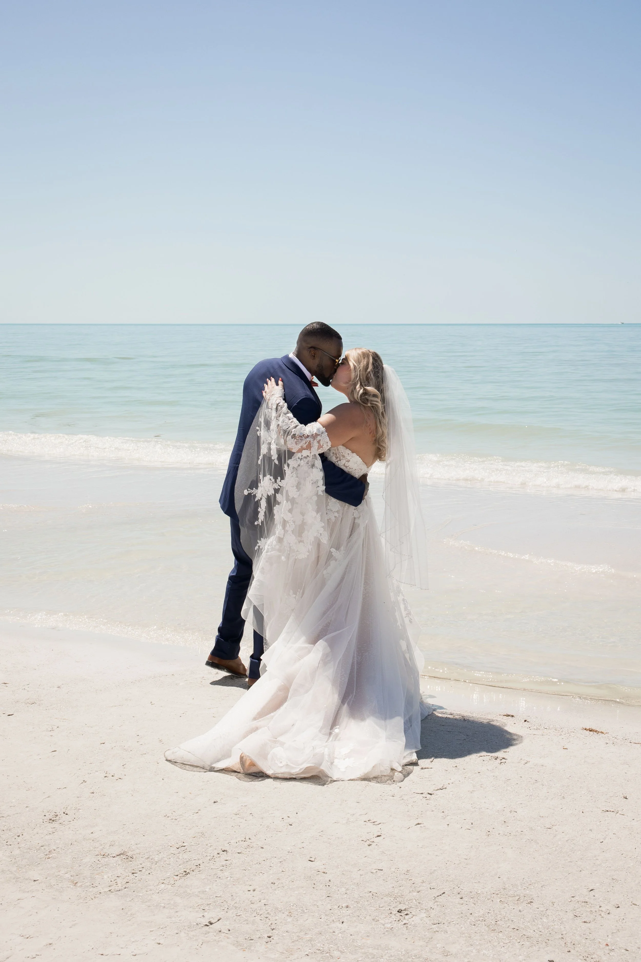 A bride and groom kissing on the beach in wedding attire, with the ocean and clear sky in the background.