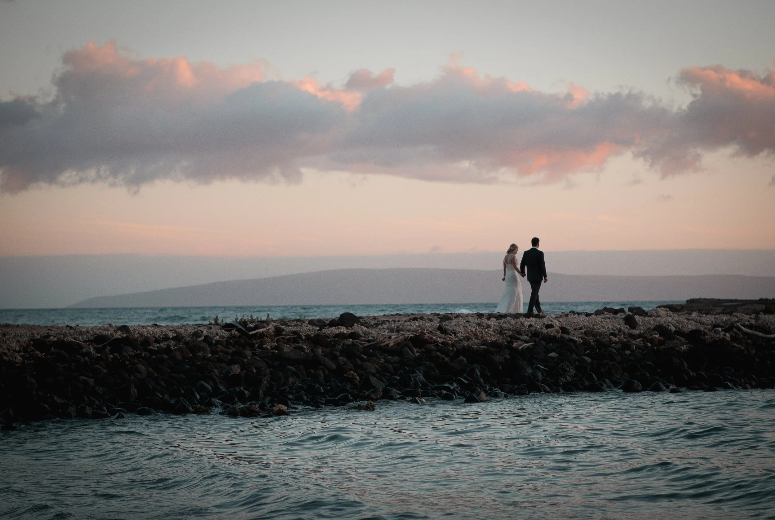 A bride and groom walking hand in hand on a rocky beach at sunset, with clouds in the sky and the ocean in the foreground In Hawaii
