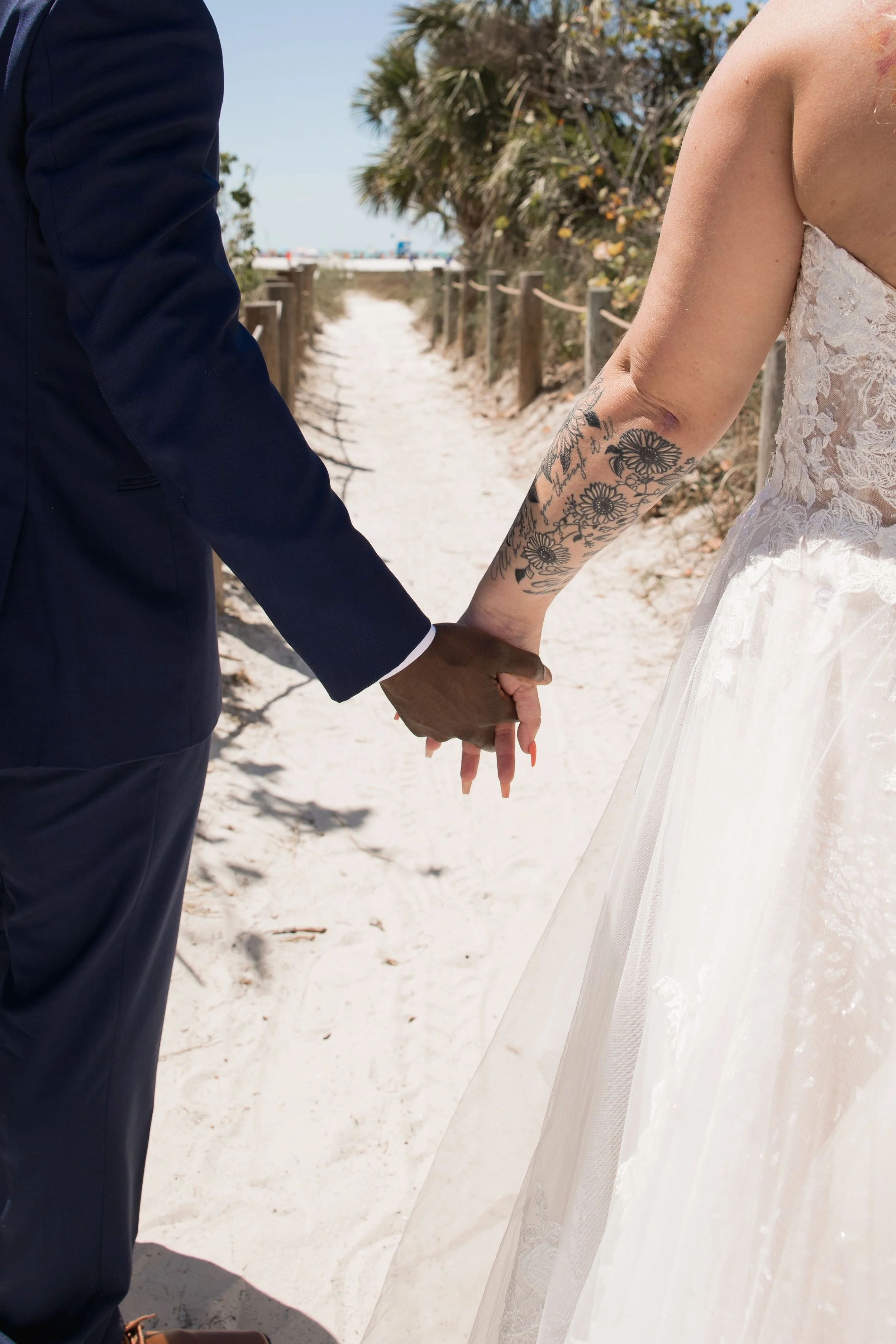 A couple holding hands on a beach walk, with the man in a navy blue suit and the woman in a lace wedding dress showing tattoos on her arm.