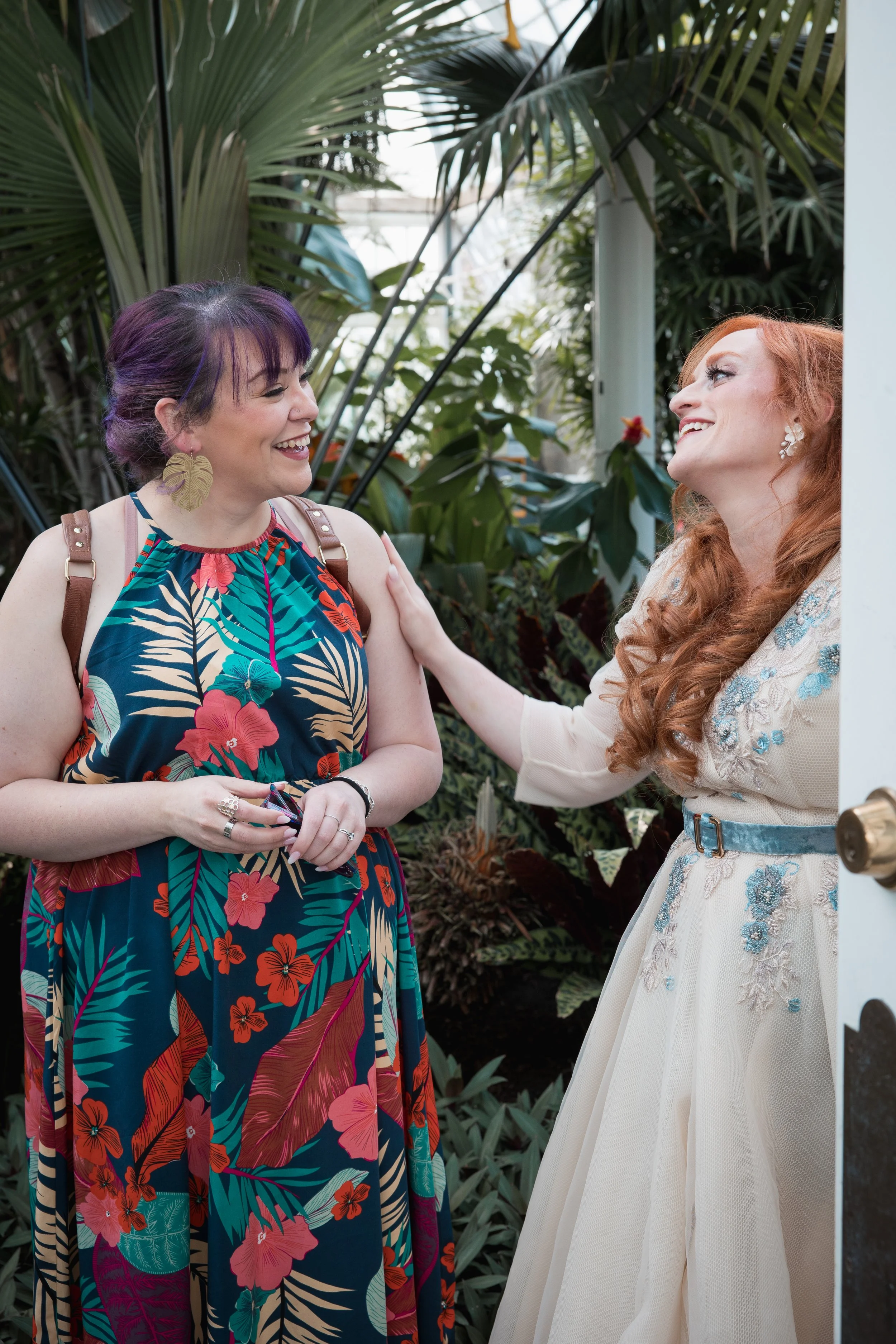 Two women are smiling and interacting inside a lush, green conservatory or tropical garden.