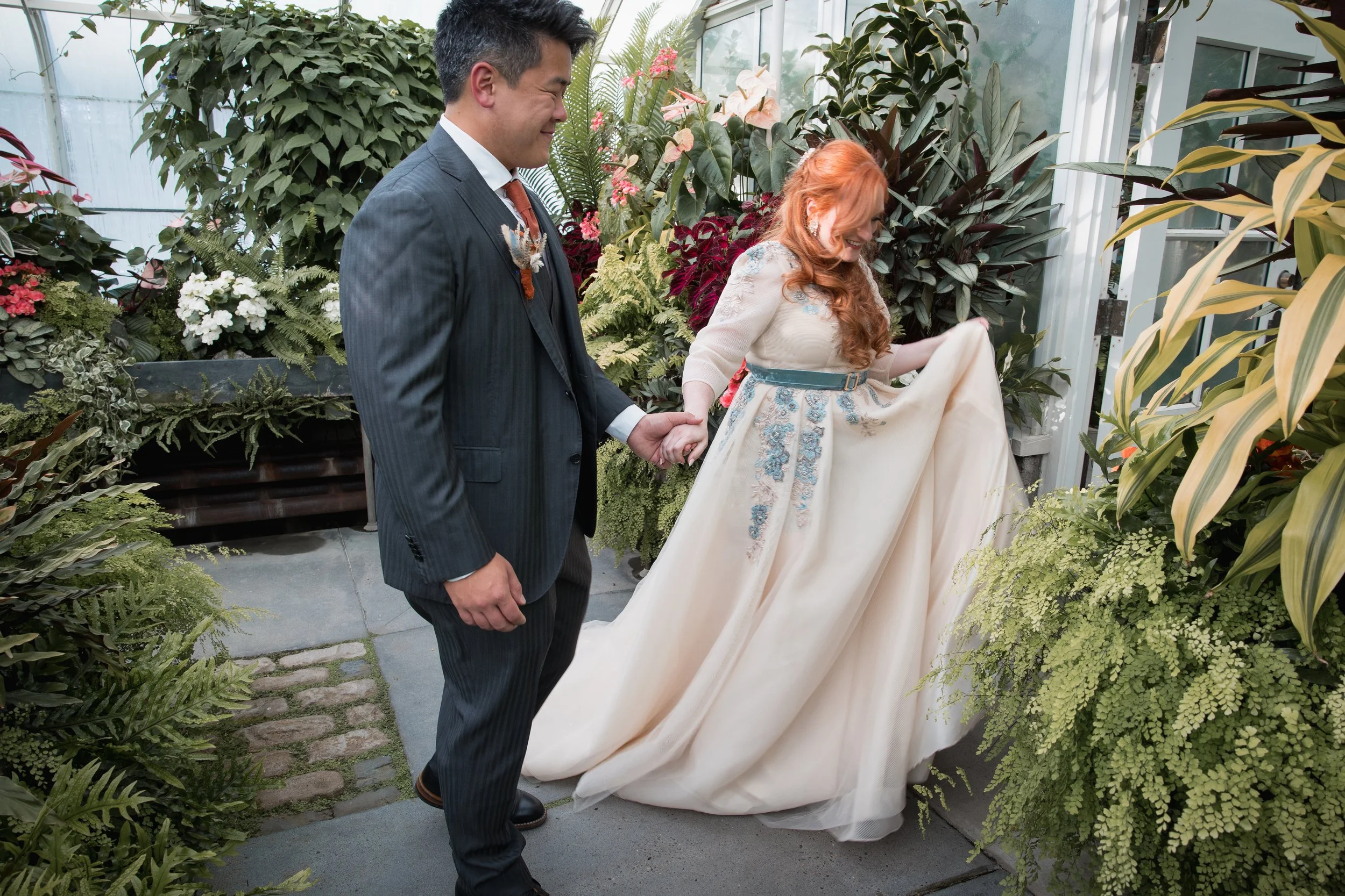 A woman in a cream-colored dress with blue embroidery lifting her dress, with a man holding her other hand, in a lush greenhouse filled with green plants and flowers in Seattle.