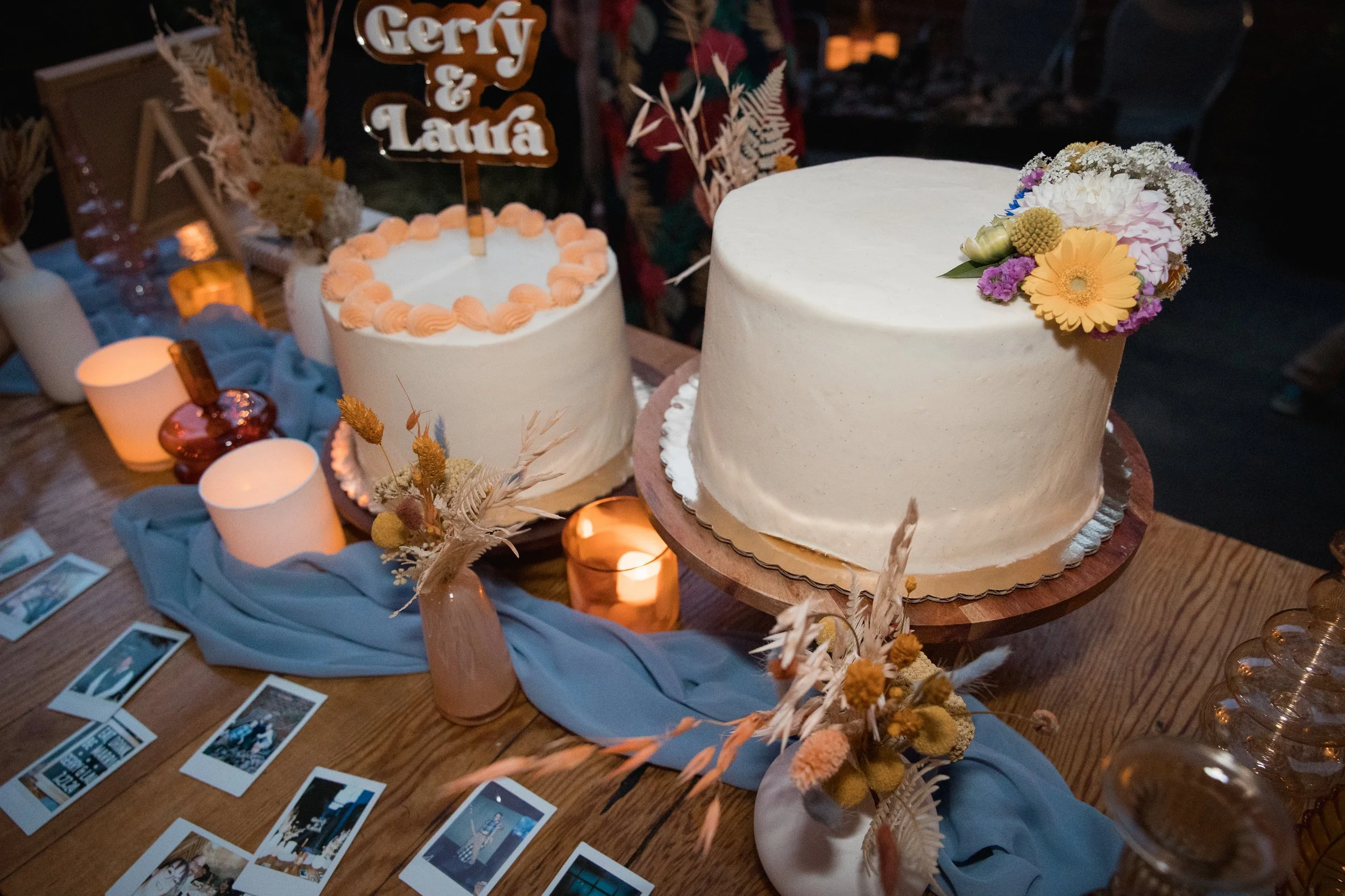 Two white frosted wedding cakes decorated with pink and white flowers and a cake topper with the names Gery and Laua, surrounded by candles, dried flowers, and photo prints on a wooden table with a blue cloth.