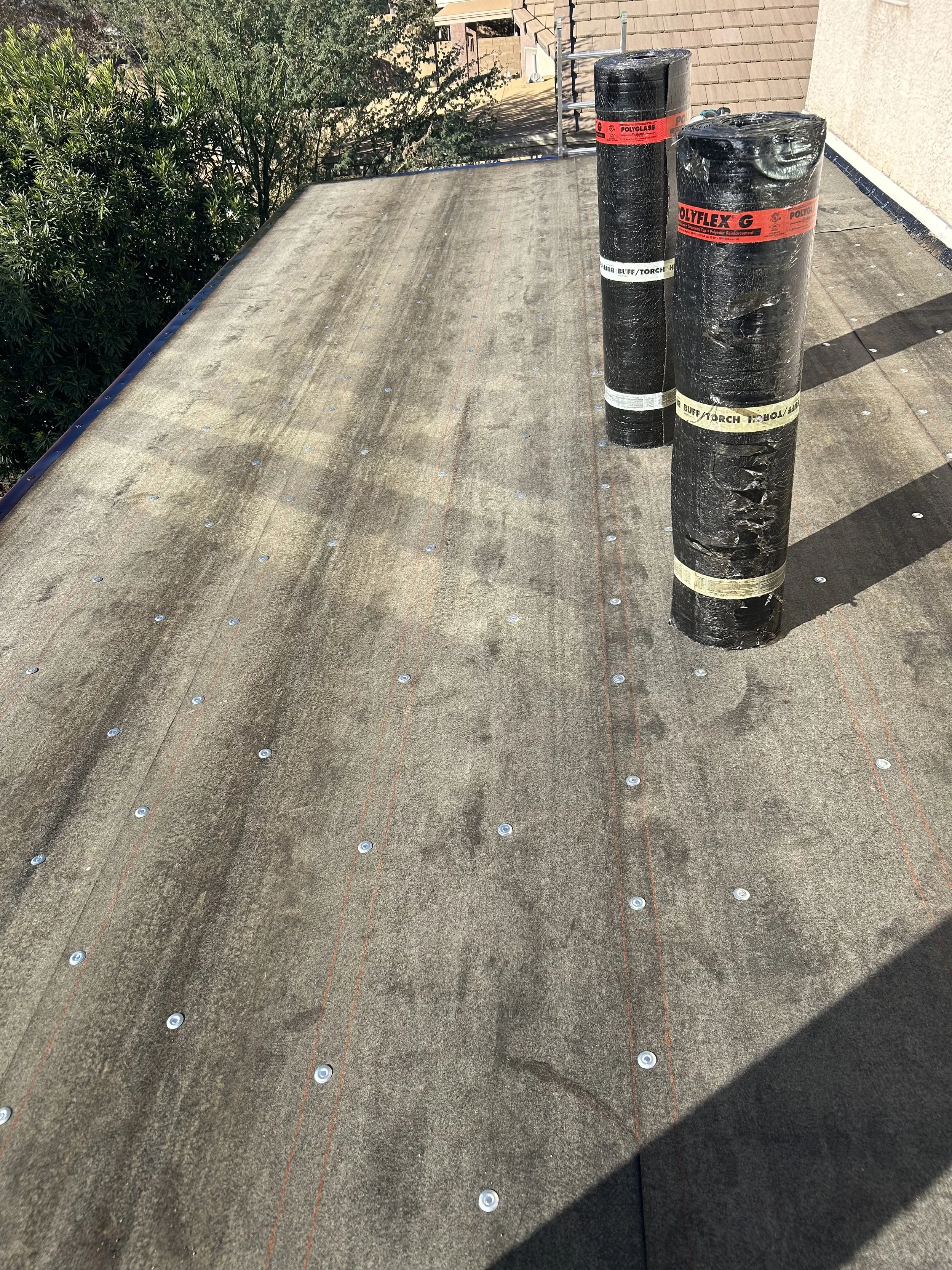 Construction workers installing a new roofing material on a house with existing shingles. The roof is partially covered with black underlayment, and tools and materials are scattered on the roof and ground.