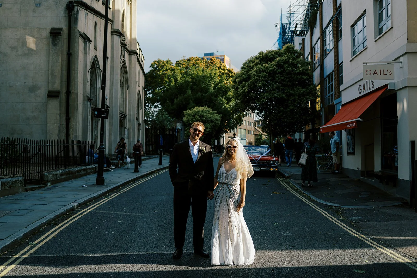 A bride and groom holding hands and smiling on a city street, with pedestrians, trees, and buildings in the background during daylight.