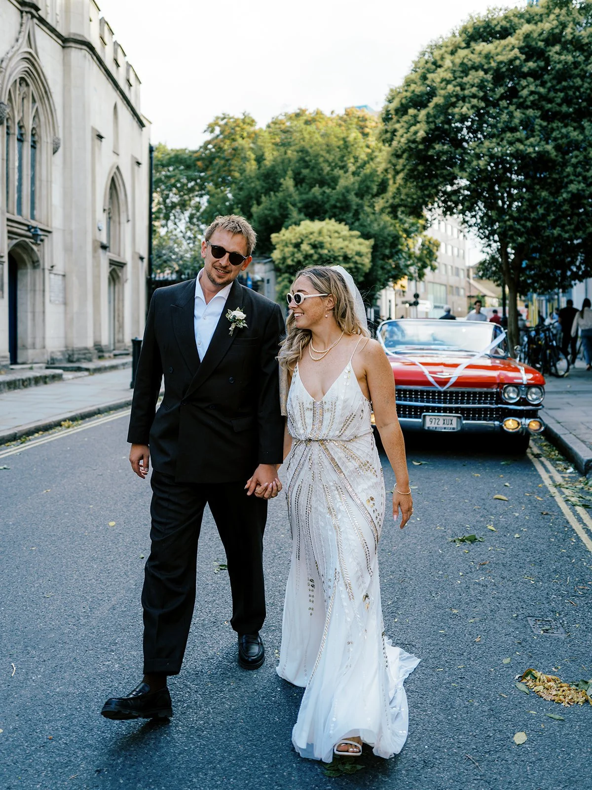 A newly married couple walks hand-in-hand down a city street, dressed in wedding attire. The groom wears a black suit with a white shirt, and the bride wears a white, beaded wedding dress and sunglasses. A vintage red car decorated with streamers is