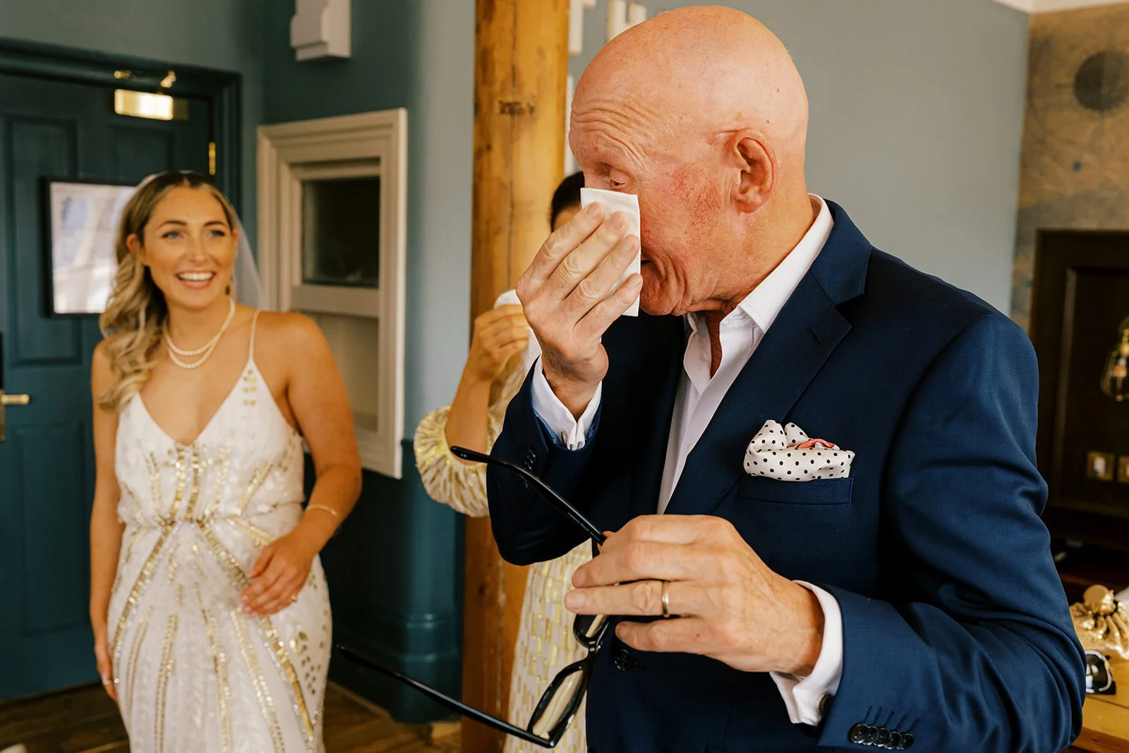 An elderly man in a navy blue suit with a polka dot pocket square wiping his eyes with a tissue while holding glasses, with a joyful woman in a white dress smiling in the background during a celebration.