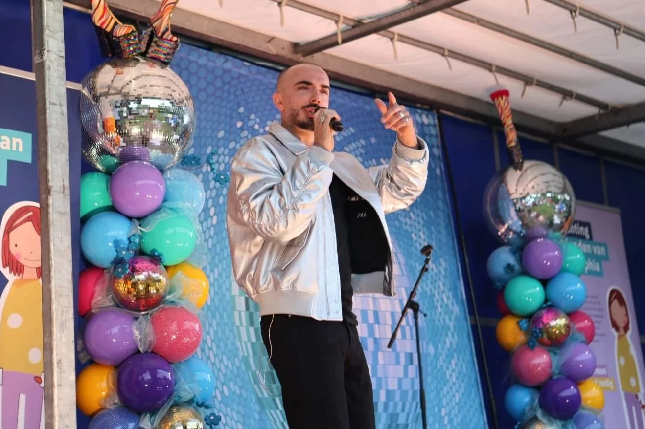 Man met een microfoon op een podium met kleurrijke ballonnen en disco ball decoraties, waarschijnlijk tijdens een optreden of evenement.