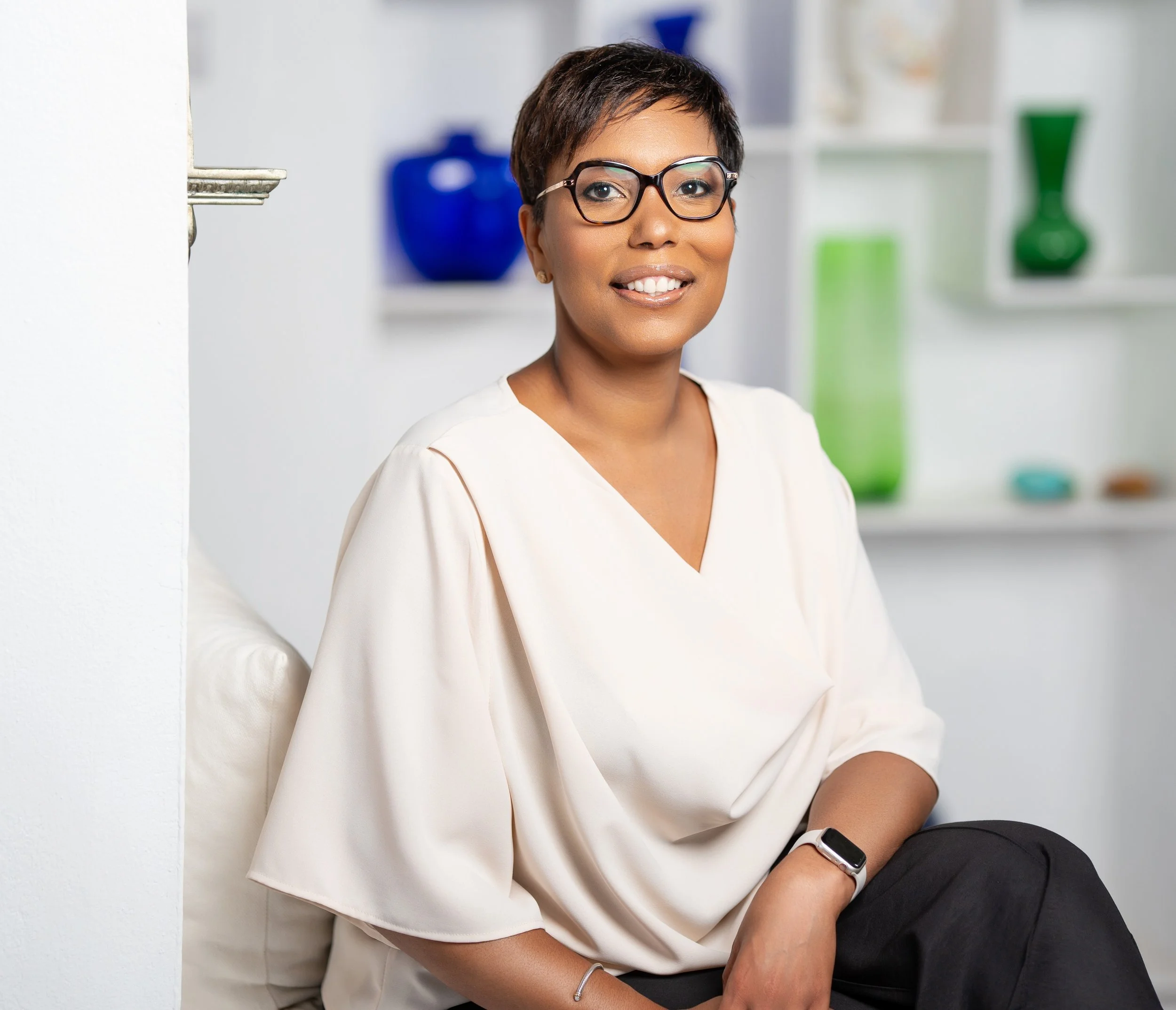 A woman with short hair, glasses, and a white blouse, sitting on a chair in a room with decorative vases on shelves behind her.