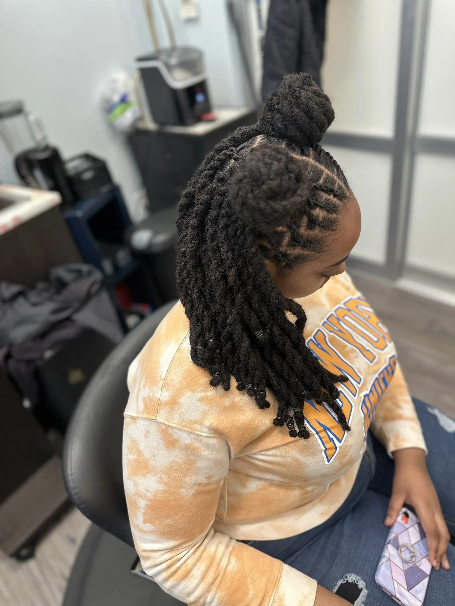 A woman with styled dreadlocks sitting in a chair, looking down at her phone, in an indoor space with office equipment and a person standing in the background.