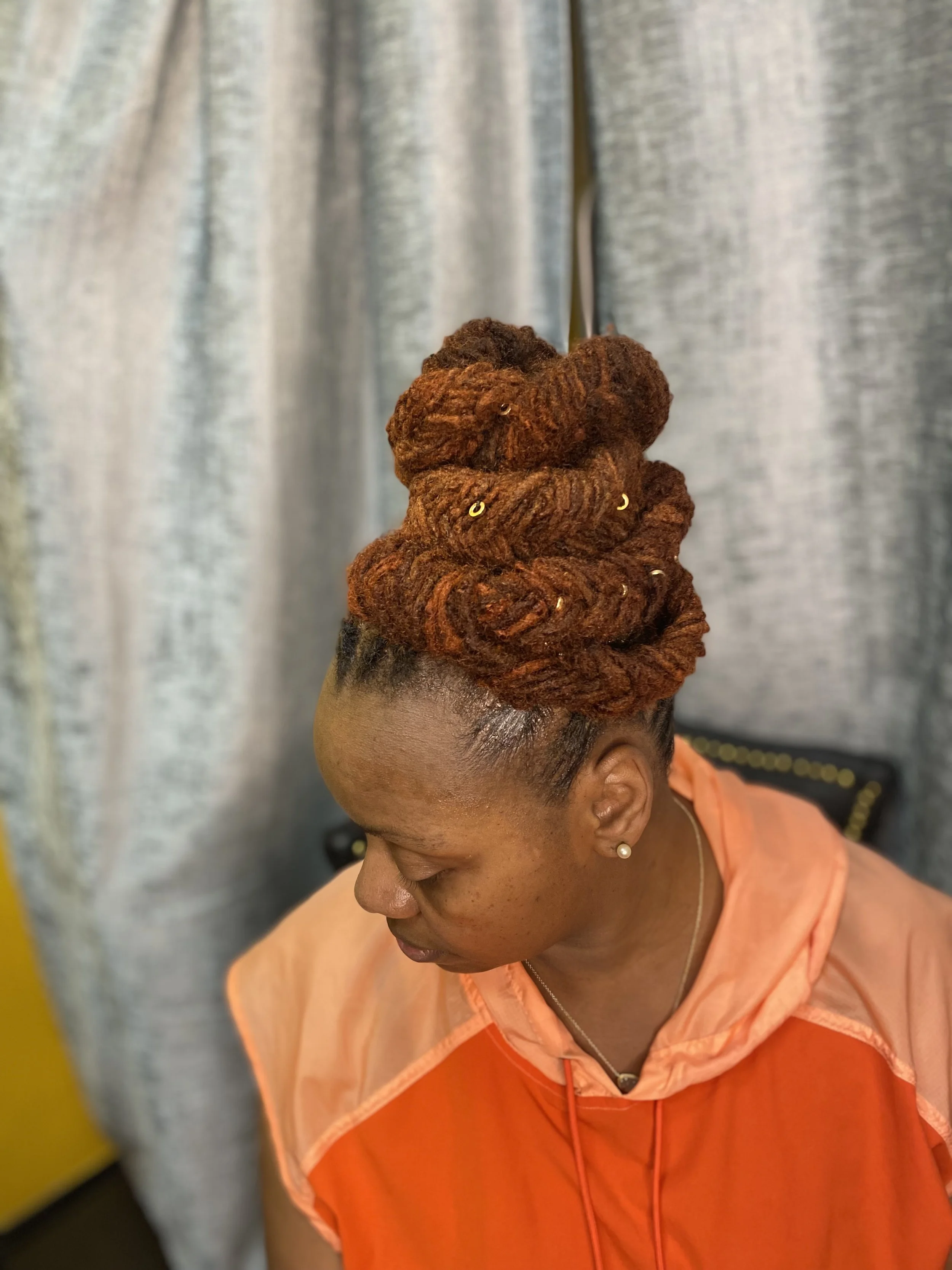 A woman with dark skin and orange dreadlocks styled in an updo, decorated with small gold rings, wearing a peach and orange jacket, looking down.