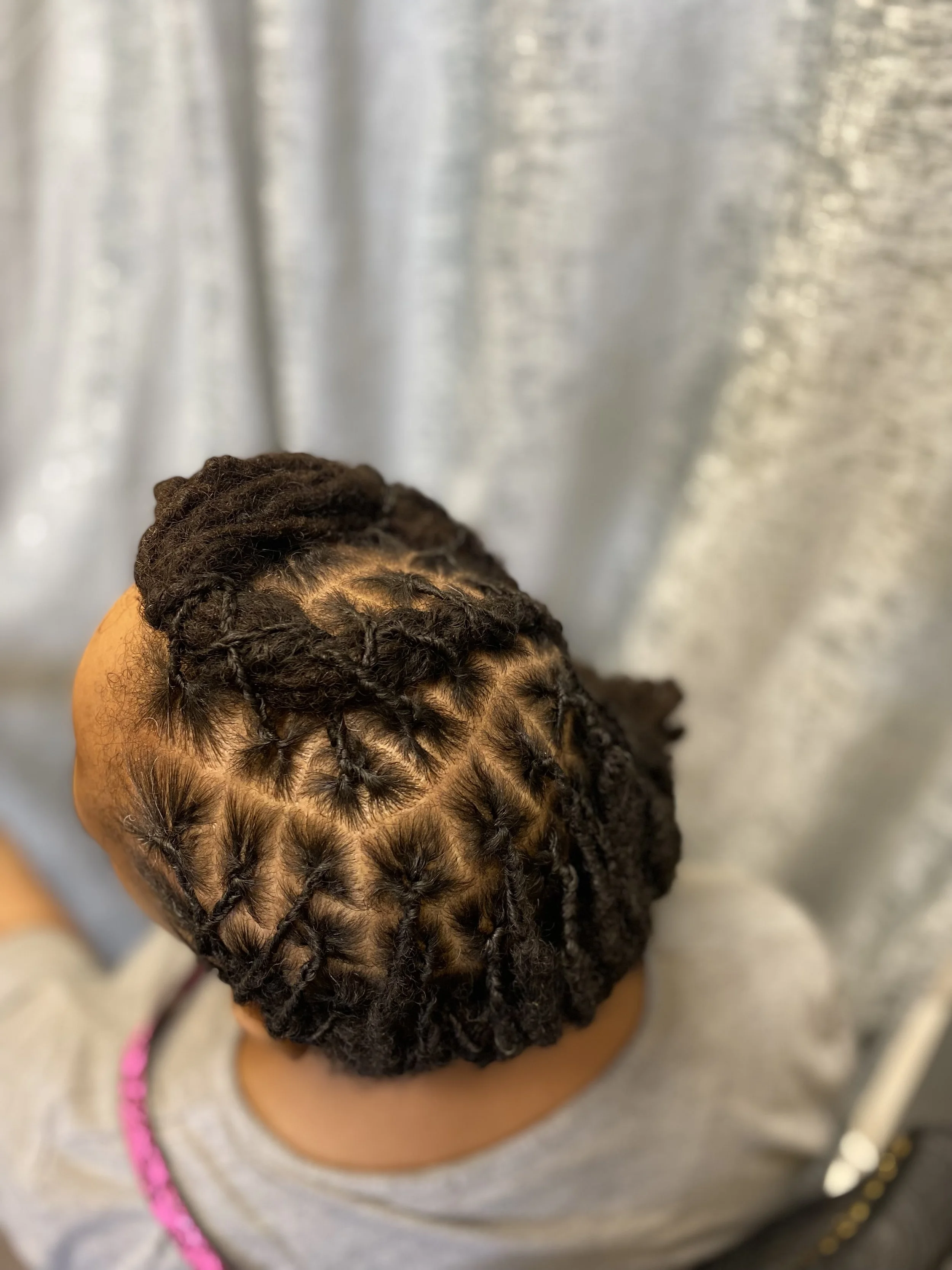 Top view of a person's head with freshly styled dreadlocks, parted to show scalp, against a blurred shiny silver curtain background.