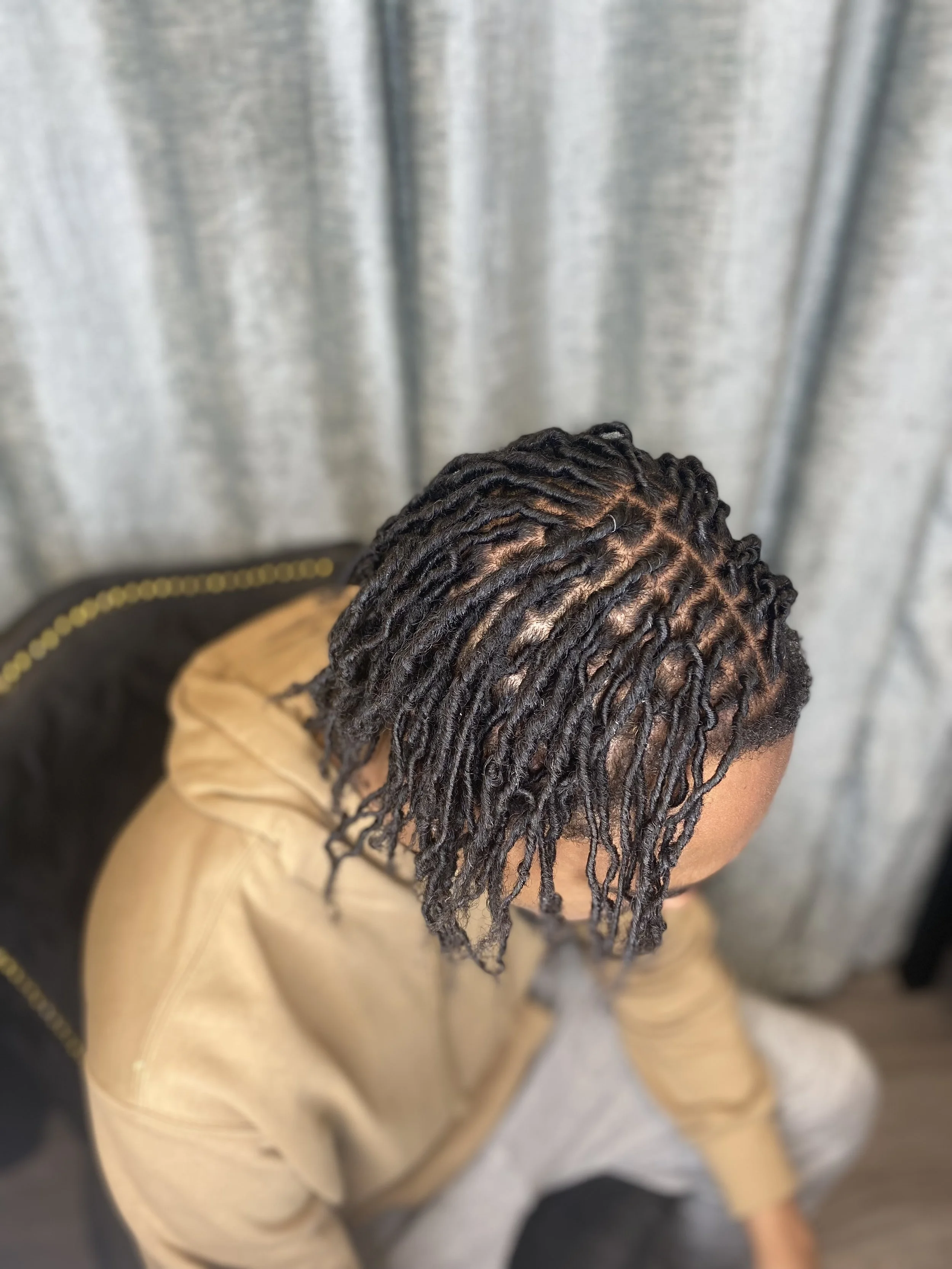 Top view of a person with freshly styled dreadlocks wearing a beige hoodie, sitting on a chair with a black backrest with gold trim, in front of a gray curtain.