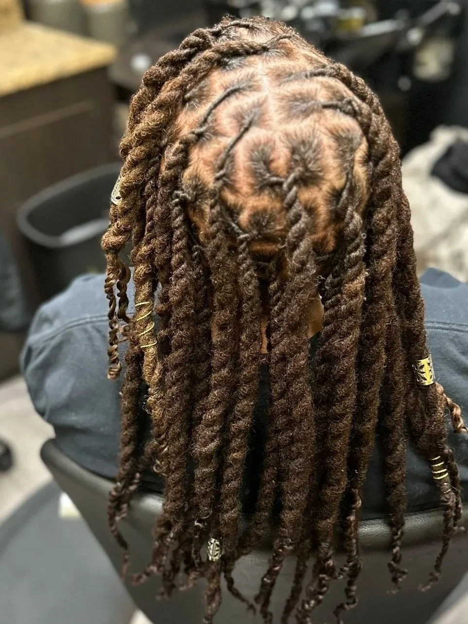 Close-up of a person with long, brown, twisted dreadlocks sitting in a salon chair, with hair sections secured with small gold-colored hair beads.
