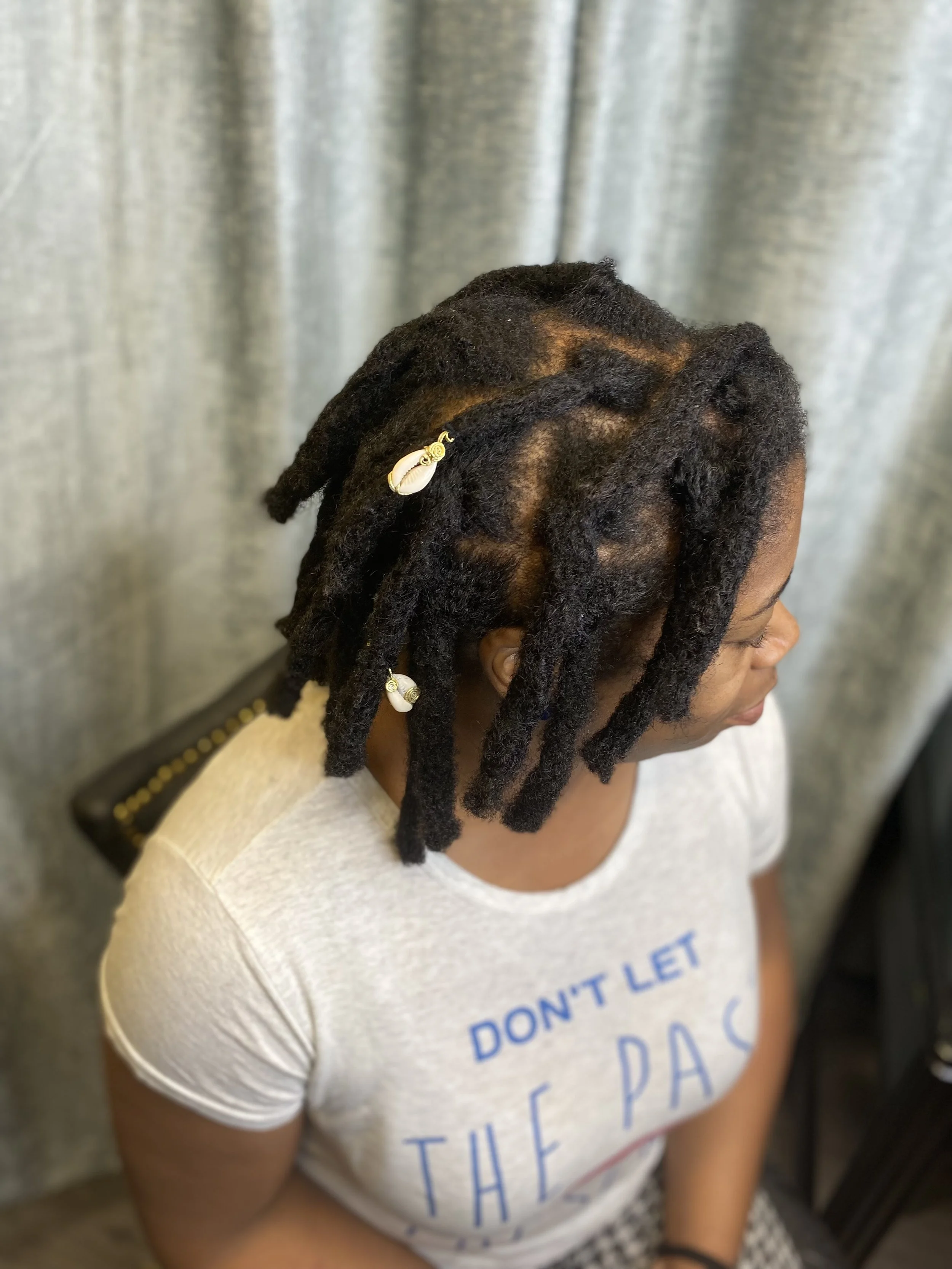 Woman with styled dreadlocks secured with shell and gold accessories, wearing a light gray T-shirt with blue text, sitting in front of a gray curtain.