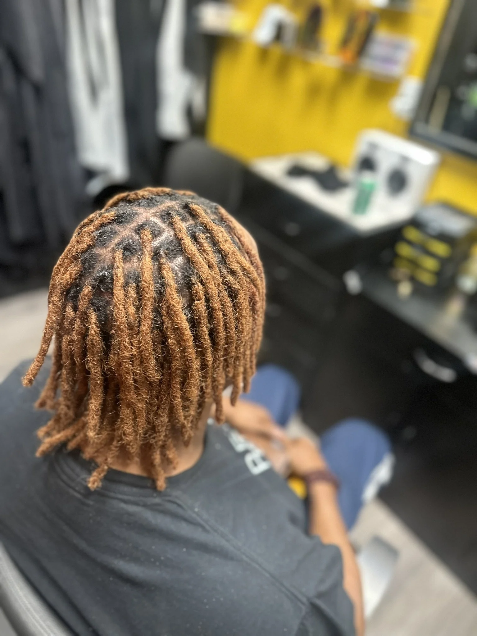 Back view of a person with blonde dreadlocks sitting in a barber or salon chair, in a space with yellow and black decor and a computer or television screen nearby.