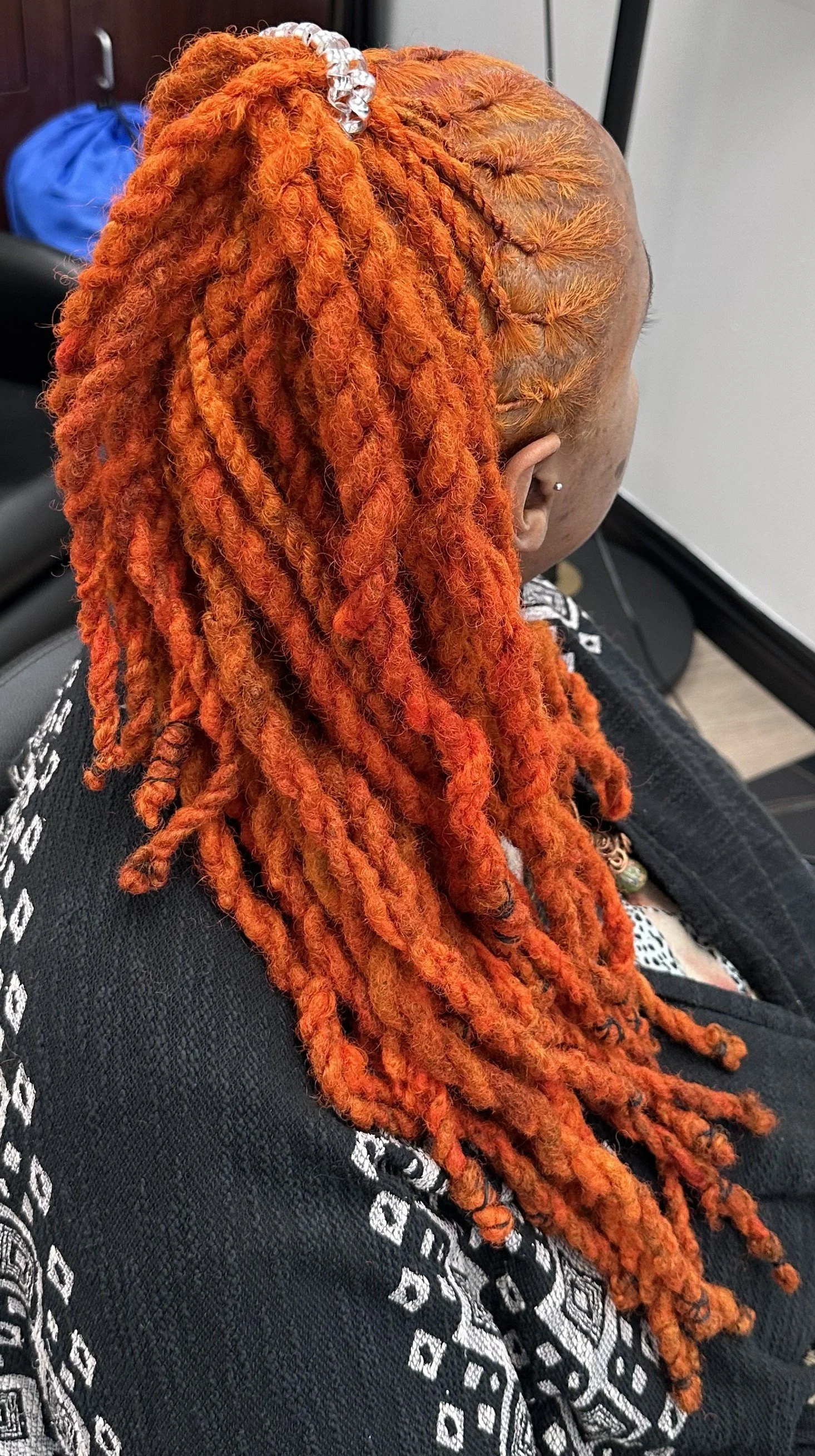 A person with bright orange dreadlocks tied back with a silver hair accessory, sitting in a chair with a black and white patterned shirt and multiple earrings.